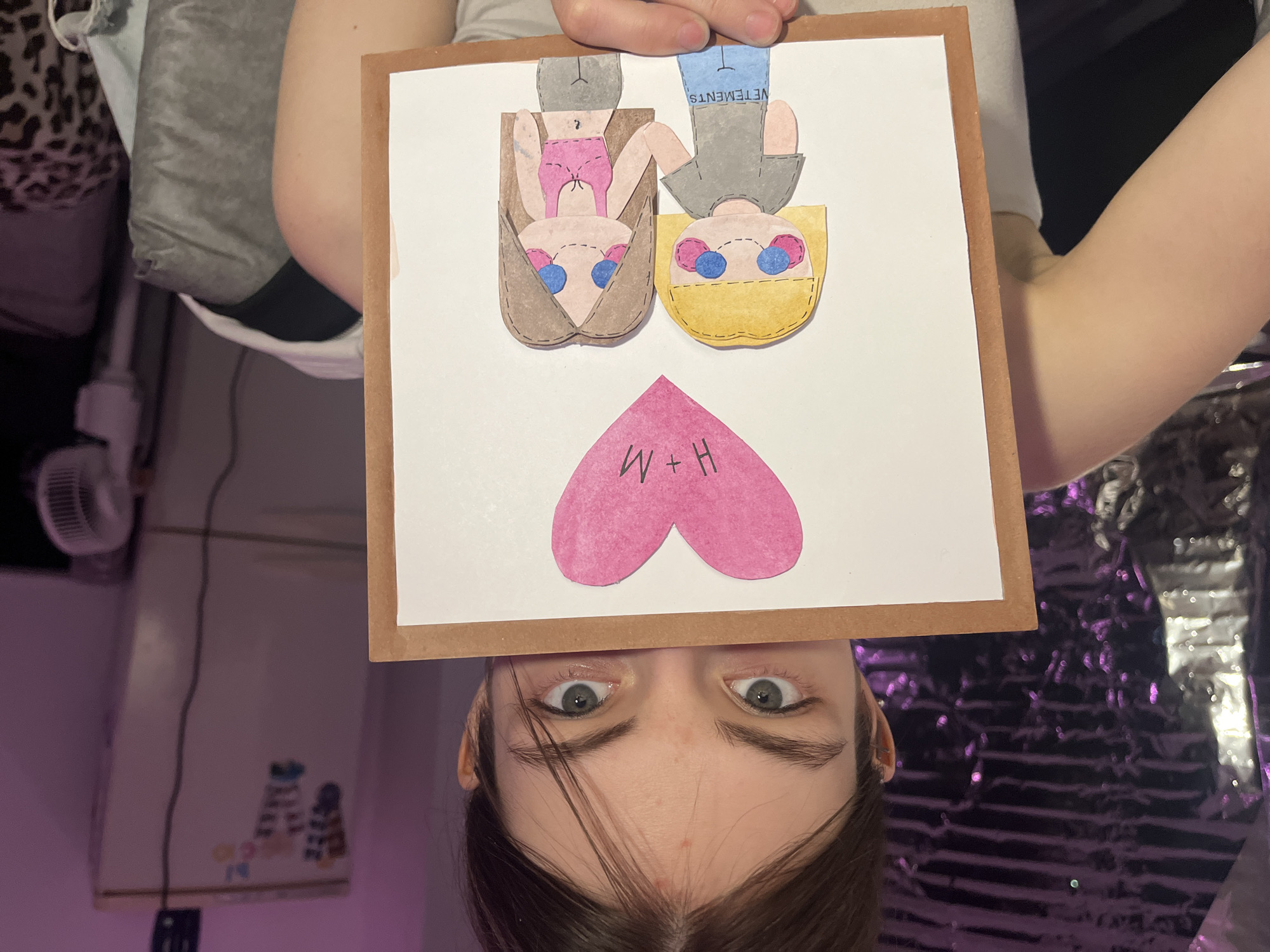 The image shows a person holding up a drawing in front of their face. The drawing is on a white square with a brown frame. The drawing features two upside-down figures, one with brown hair and clothing and the other with blonde hair and grey clothing labeled "VETEMENTS." A pink heart with the letters "W+H" is below the figures. The person holding the drawing has brown hair, light skin, and is looking up at the camera. The background is blurred and features a silver, textured material and a white air conditioner. The location is difficult to ascertain from the image and cannot be narrowed to Saint Petersburg, Russia.