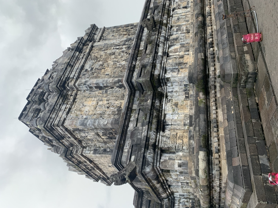 The image shows the exterior of a large stone temple, likely a Buddhist temple, located in Mungkid, Indonesia, at the coordinates -7.605 latitude and 110.23 longitude. The temple is made of weathered gray stone blocks and is covered in moss and lichen. There are several decorative carvings on the temple walls. The temple is surrounded by a paved courtyard, with a pink propane tank in the foreground. The sky is overcast.
