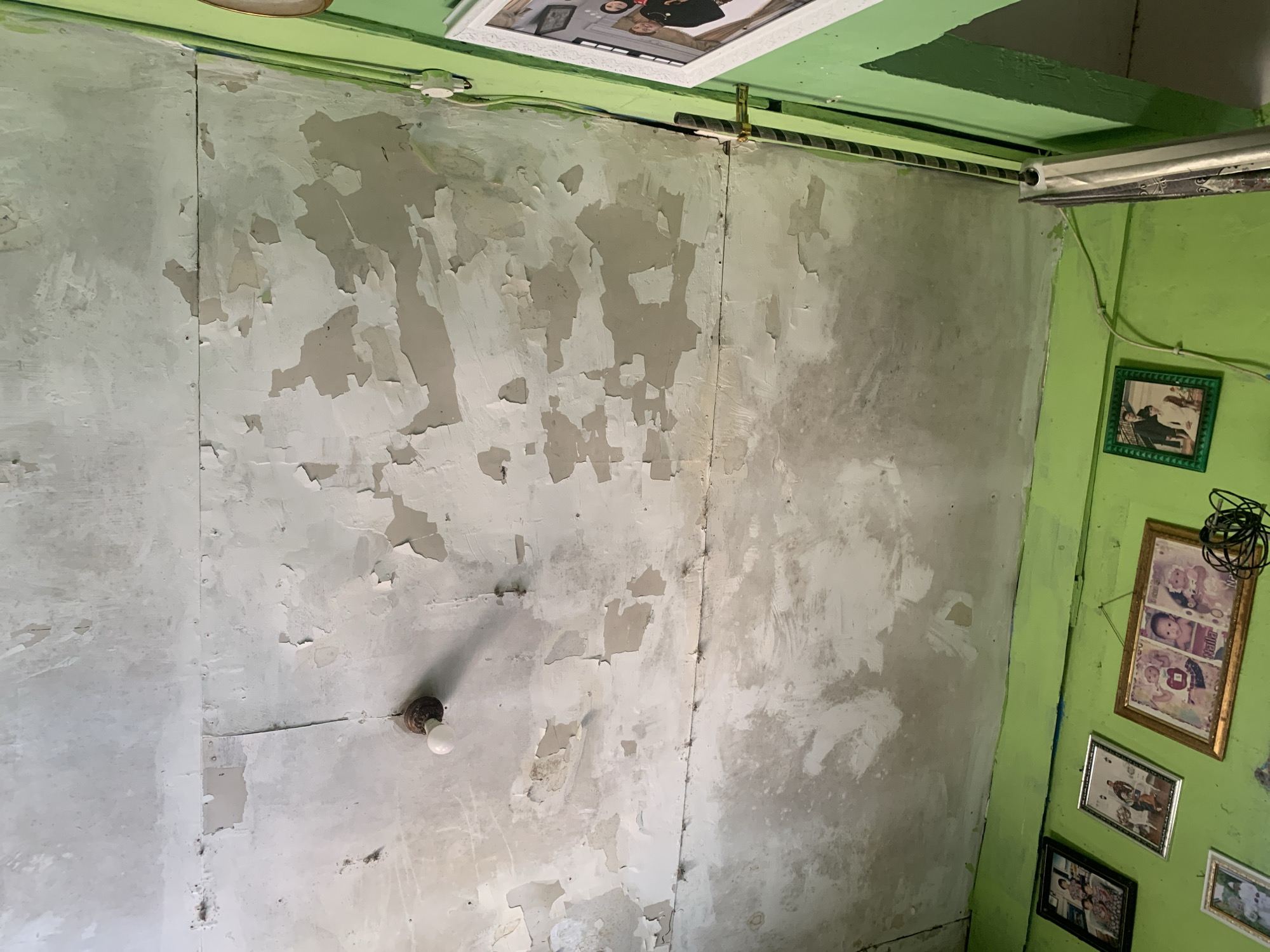 The image depicts a partially renovated or damaged interior wall with peeling paint, located in Palembang, Indonesia. The wall is a light gray and shows significant areas where the paint has flaked off, revealing a rougher surface beneath. A single bare lightbulb is attached to the wall near the center. On the right side of the image, a bright green painted wall is visible, adorned with several framed pictures. The frames are of various sizes and designs, with one prominently displaying a golden frame with what appear to be photographs of children. The ceiling is also painted green and has a white framed picture hanging from it. Electrical conduits and wires are visible along the top edge of the wall and ceiling. The overall impression is one of disrepair and ongoing work or neglect within a residential or commercial space.