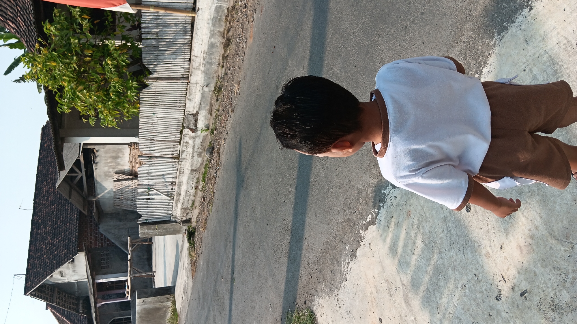 A young boy stands on a concrete path, facing away from the camera, looking towards a street in Sonobekel, Indonesia. He is wearing a white t-shirt with brown trim and brown shorts. The street is paved and asphalt. To the right of the street, a wooden fence made of bamboo stalks encloses a property. Behind the fence, a house with a tiled roof and concrete walls is visible, with a small tree growing nearby. The sky is clear and bright.