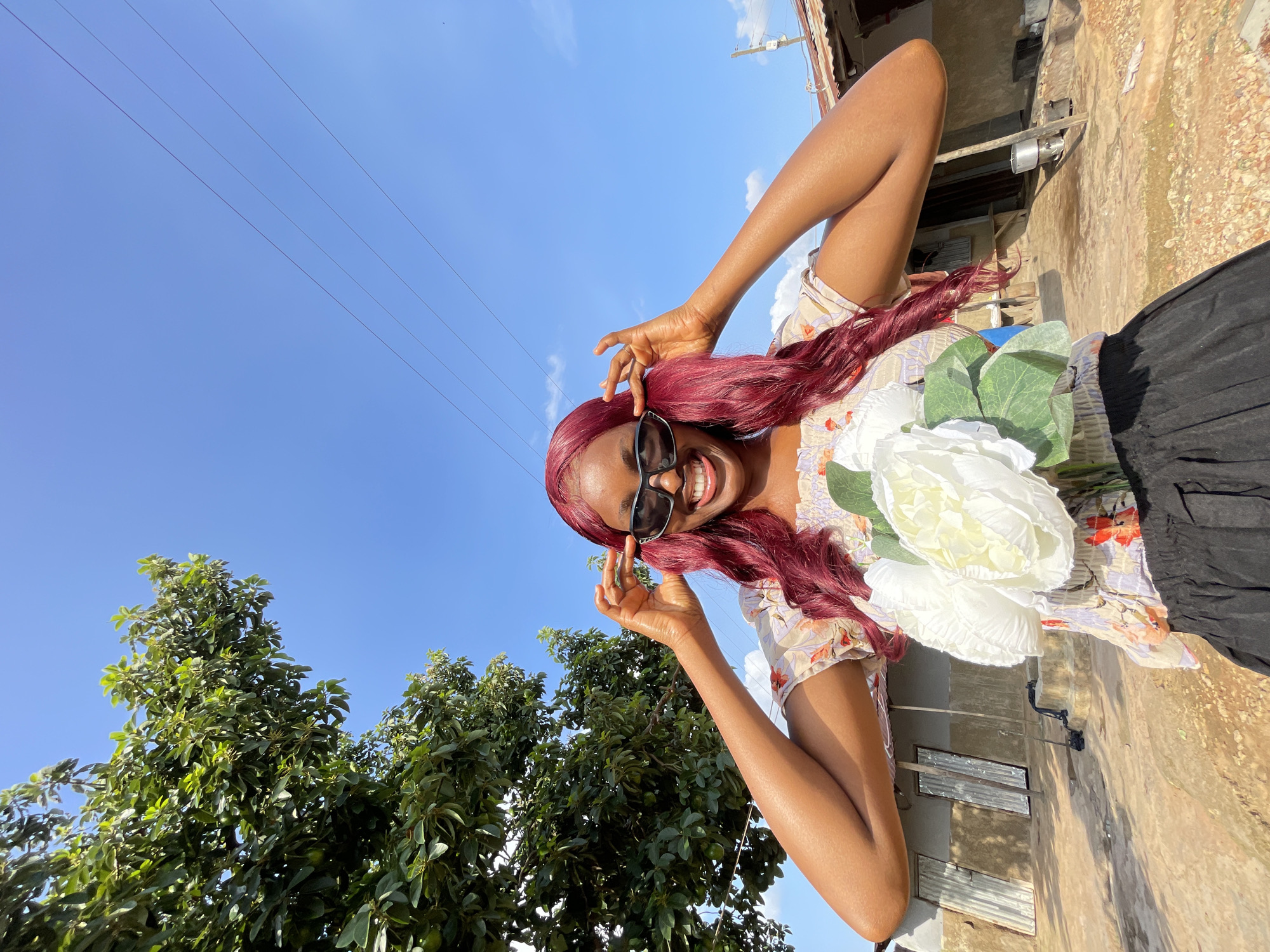 A young Black woman with long, dark red hair and sunglasses smiles broadly at the camera, holding a bouquet of white artificial flowers. She is wearing a light-colored, floral print off-the-shoulder top and a black skirt. She is outdoors in a rural setting, with a lush green tree to her left and a rustic building with corrugated metal roofs in the background. The ground is dirt and appears dry. The sky is bright blue with a few scattered clouds, and power lines are visible stretching across it. The scene suggests a warm, sunny day in a village outside of Jos, Nigeria.