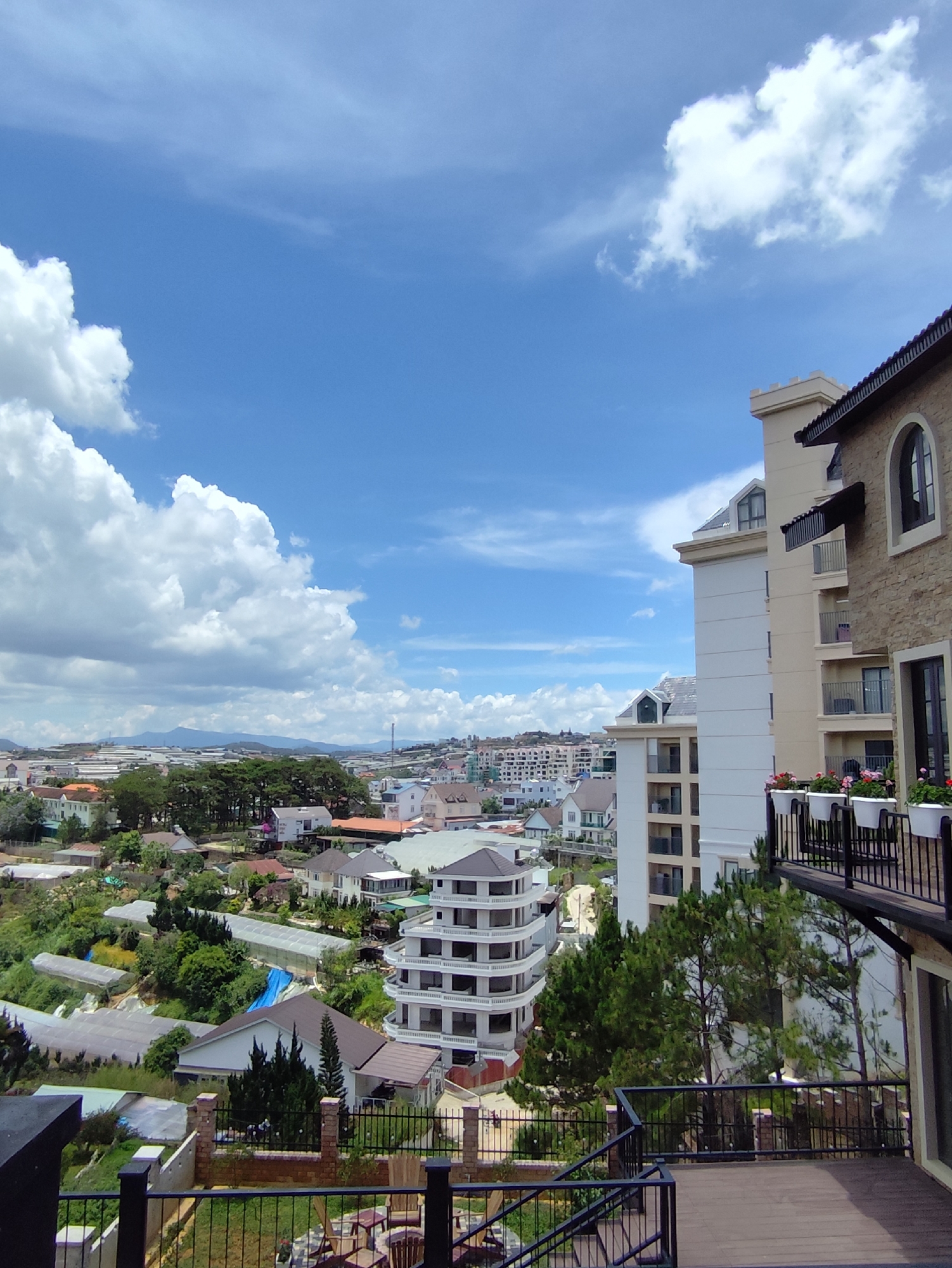 This is an eye-level, outdoor daytime shot taken from a balcony or patio, showcasing a view of Xuân Trường - Đà Lạt, Vietnam.
The foreground has a wooden deck with black metal railings. Chairs and landscaping are partially visible.
The midground features residential buildings, a multi-story, curved white building, and a series of greenhouses. Trees are interspersed. The urban landscape extends toward the horizon, where hills and distant mountains are visible.
The background consists of a vibrant blue sky filled with cumulus clouds.