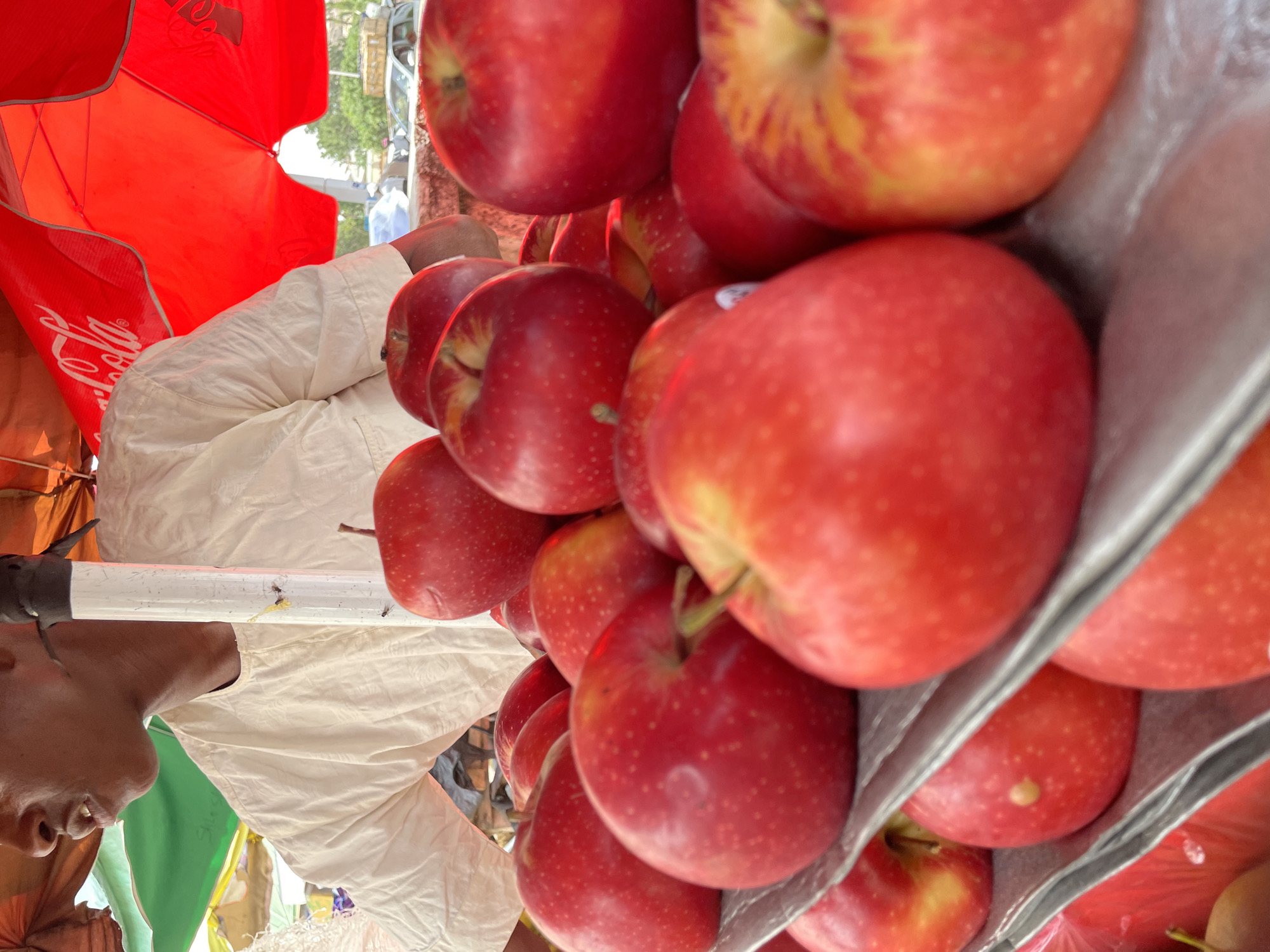 A collection of red apples in a grey plastic container is visible. A person, identifiable by their face and arms, is beside the container. A red Coca-Cola branded umbrella is partially visible overhead. The image appears to be taken at an upward angle. The setting is likely a market or roadside stall in Jos, Nigeria, due to the presence of the apples and the stall-like structure.