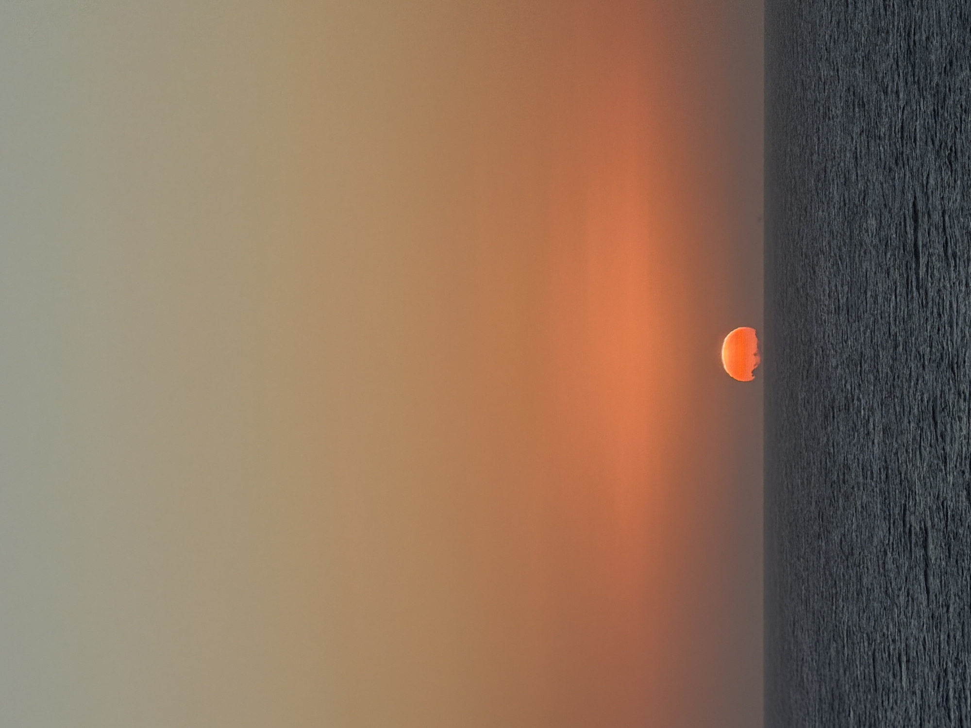 The image depicts a sunset over a body of water, possibly an ocean or a large lake. The sky transitions from a light grey-beige to a warm orange hue near the horizon. A bright orange disc, representing the sun, is partially visible just above the water line. The water itself appears dark and textured, suggesting a choppy surface or perhaps detailed rendering of the water's surface. No specific location or city can be identified based solely on the visual elements in the image. There are no people visible in the scene.