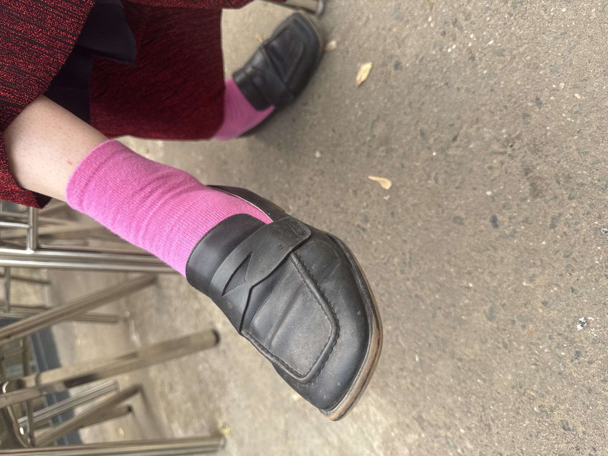 The image shows a person's legs and feet clad in pink socks and black loafers. The person is seated, with metal chair legs visible on the left side of the frame. The surface beneath the chair and feet is concrete. The photo appears to be taken outdoors. There is no evidence to determine the exact location of the photo is Moscow, Russia.