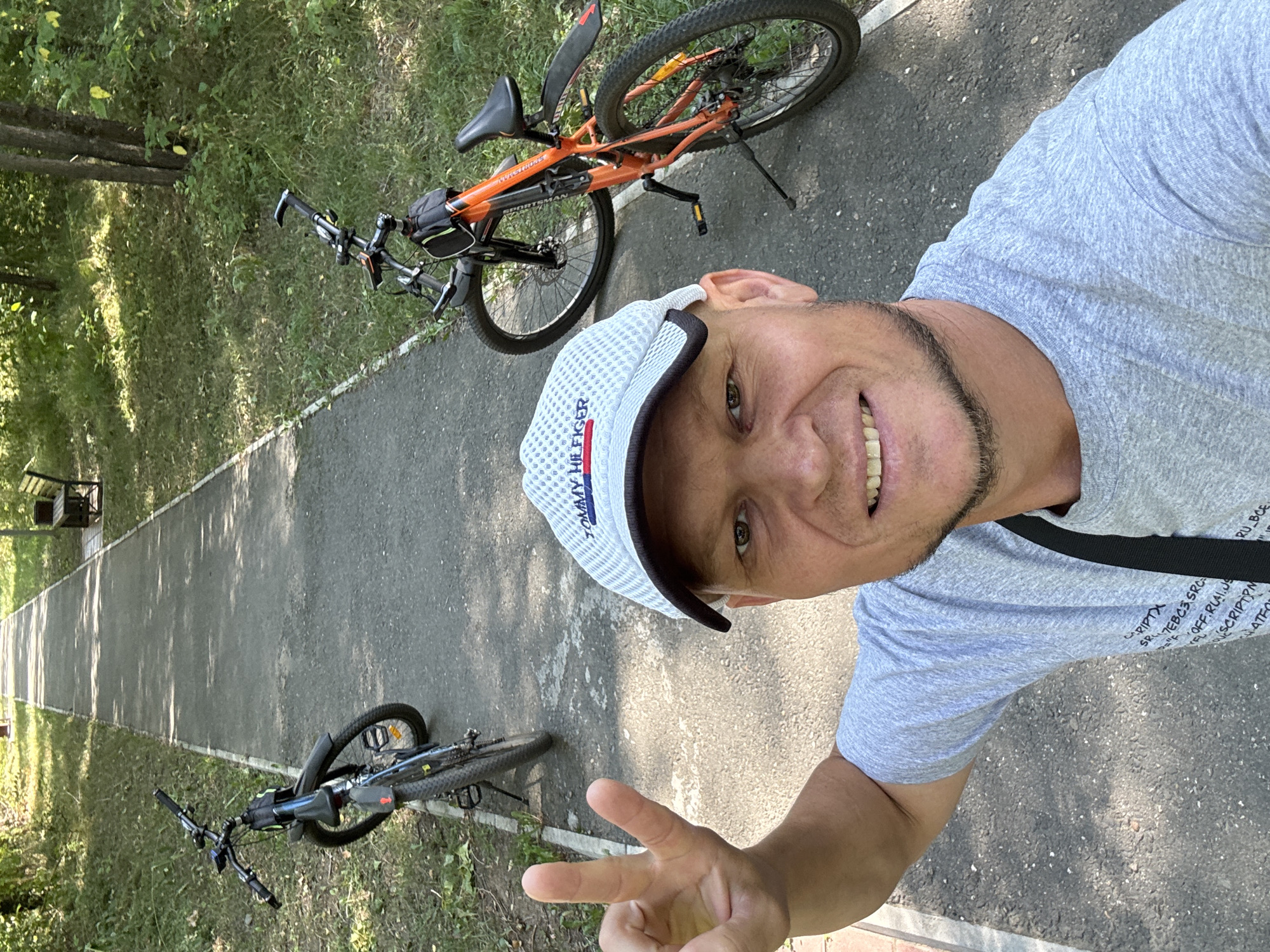 A man in a gray t-shirt and a Tommy Hilfiger baseball cap takes a selfie outdoors, with two bicycles parked on a paved path behind him. He is smiling and holding up two fingers in a peace sign with his left hand. The path is lined with grass and trees, and a wooden bench is visible in the background. The scene appears to be a park or recreational area in the city of Oskemen, Kazakhstan.