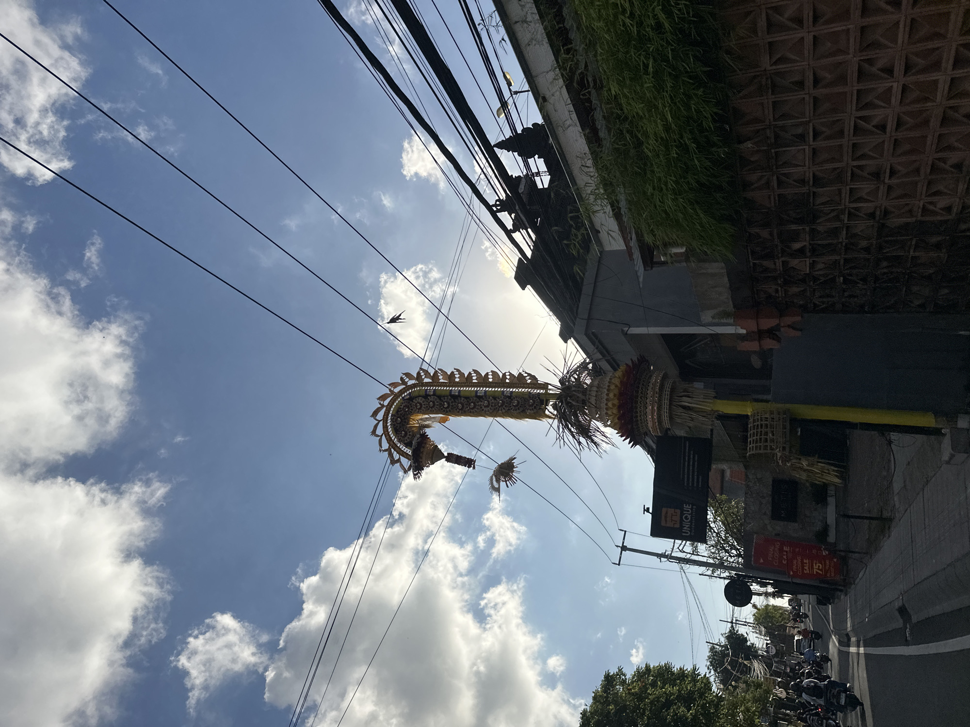 A low-angle outdoor shot captures a street scene in North Kuta, Indonesia, under a blue sky with scattered clouds. A tall, ornate golden decoration, possibly a Balinese ceremonial offering, dominates the foreground, reaching towards the sky and silhouetted against the sun. Below it, a street with a white-lined asphalt road is lined with buildings and a sidewalk. A few trees are visible on the left. Several motorbikes are on the road, with people riding them. Buildings on the right side of the street have their upper portions visible, some adorned with traditional architectural elements. Power lines crisscross the sky above the buildings and the street. A sign on one building reads "UNIQUE Outdoorliving." Another sign indicates "FINAL CLOSING SALE 75%." The overall impression is a bustling urban street with cultural elements.