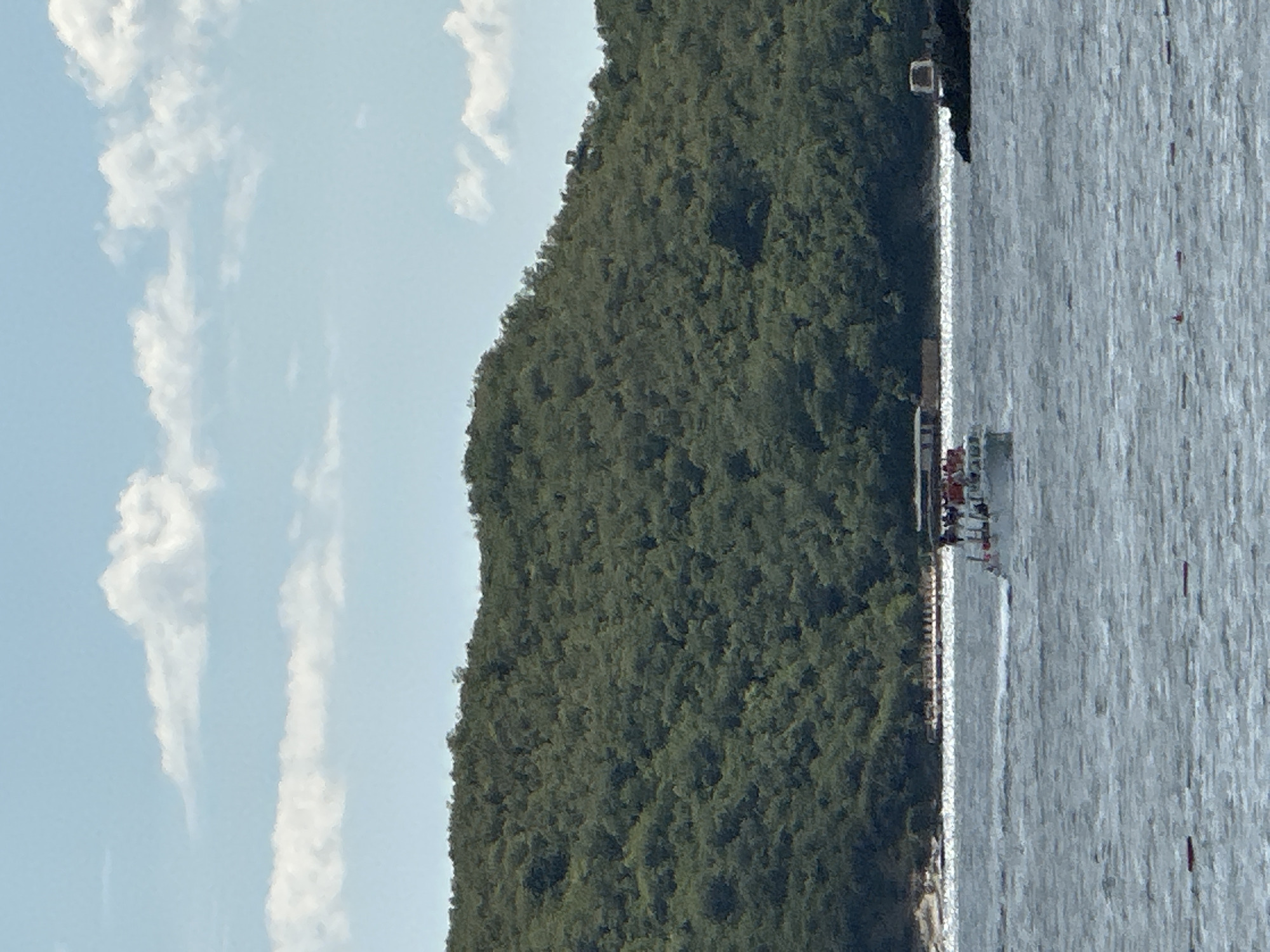 This is a full shot image of a boat in the water with a green, tree-covered hill in the background. The sky is blue with scattered white clouds. The boat is white and has several people on it. The water is choppy. There are also some buoys in the water. The image appears to be taken on a sunny day. The location is the coast of Boreti, Montenegro.