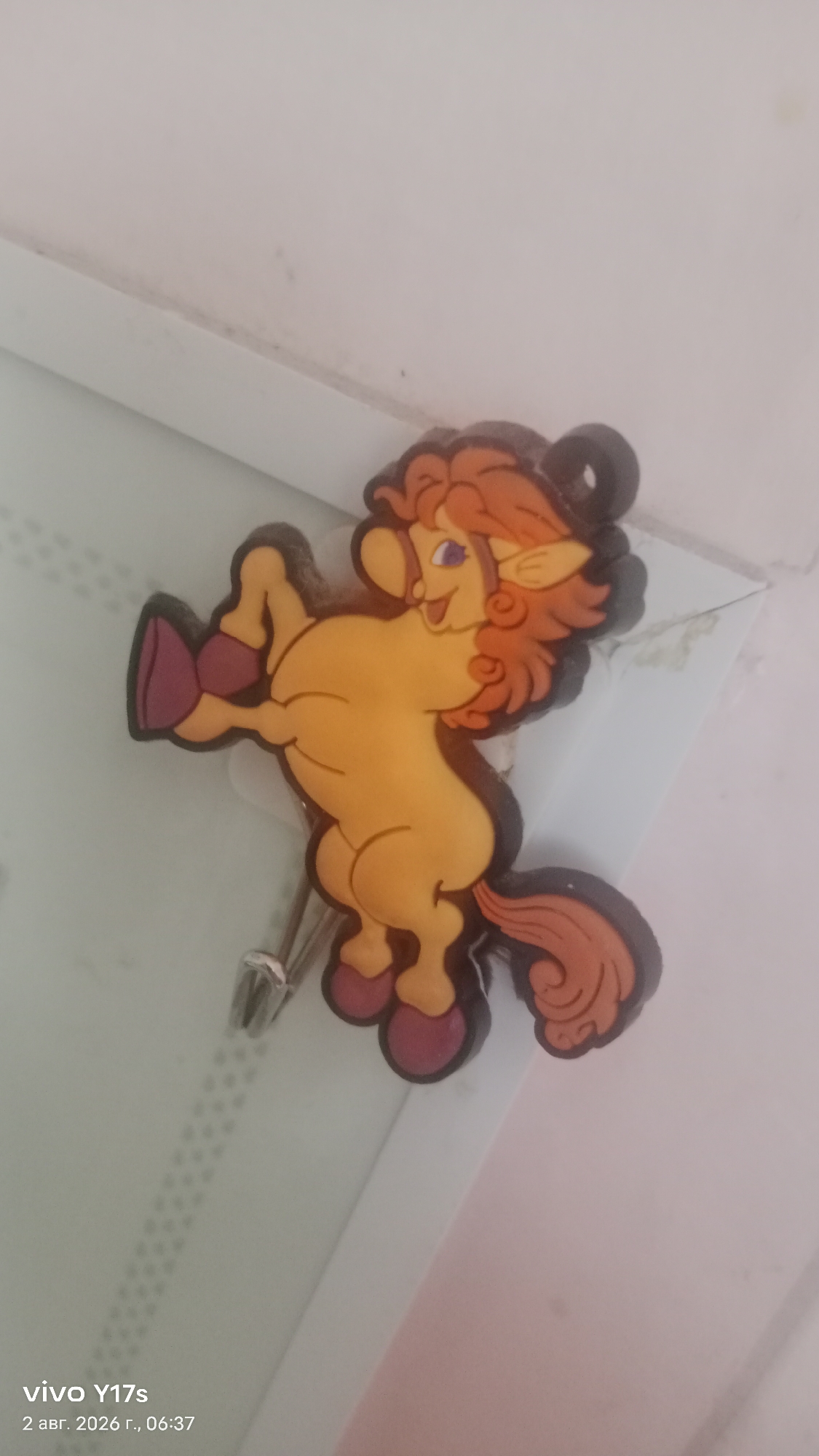 A cartoonish, anthropomorphic horse figurine, predominantly yellow with orange hair and a dark brown outline, is attached to a silver hook on a light-colored, textured surface. The horse has purple hooves, a smiling face with blue eyes, and its body is positioned as if rearing up. The background consists of a light-colored wall, possibly a closet door, with some subtle patterns visible. The bottom left of the image displays a "vivo Y17s" watermark and the date "2 авг. 2026 г., 06:37". The scene appears to be indoors, in a residential setting. No specific location within Rudny, Kazakhstan, is discernible from the provided image.