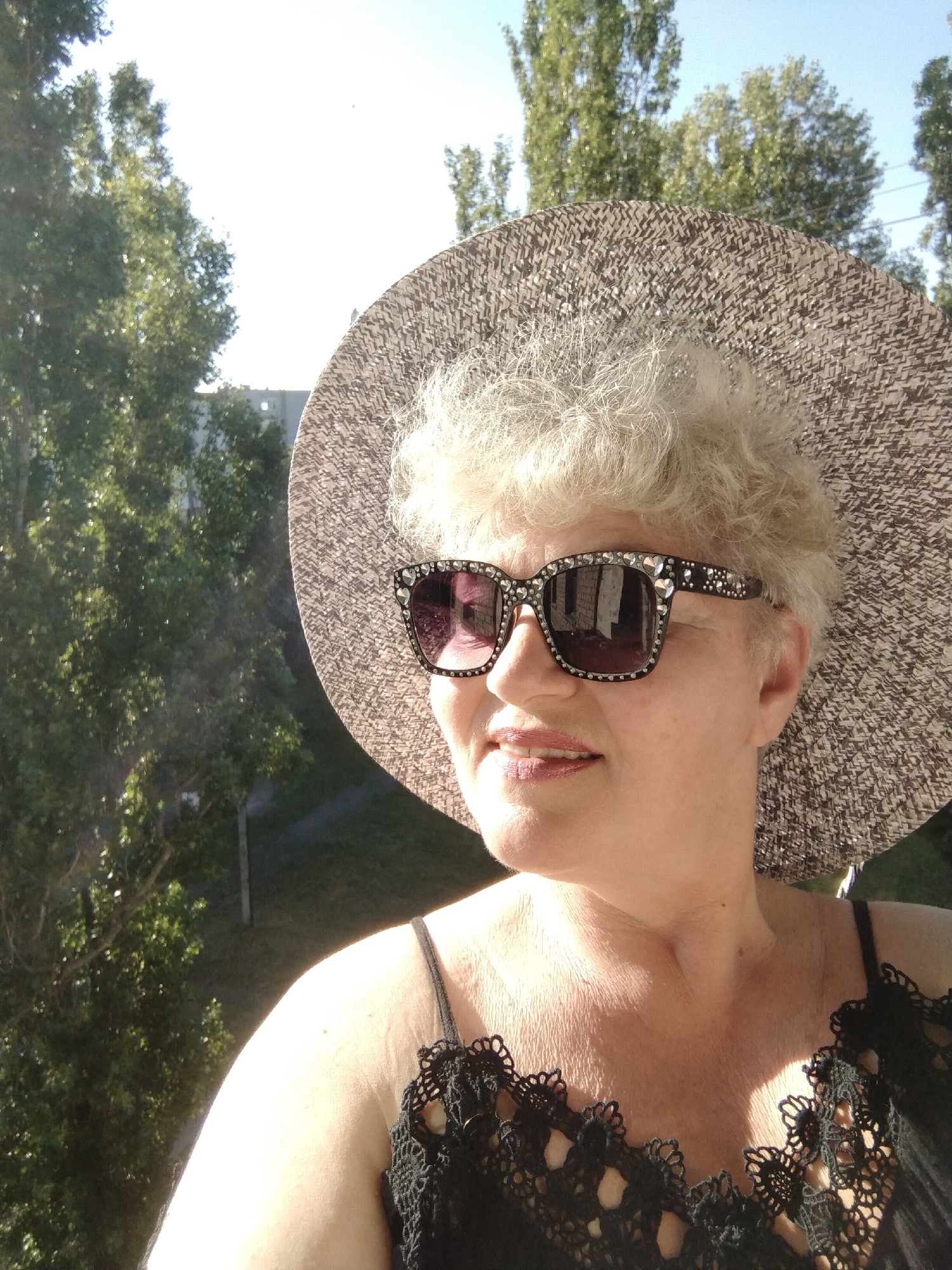 The image captures a woman outdoors in bright sunlight. She has short, curly, light-colored hair and is wearing a wide-brimmed straw hat with a woven brown and beige pattern. Her face is turned slightly to her left, and she has a gentle smile. She wears stylish sunglasses with dark lenses, the black frames of which are adorned with numerous small, sparkling embellishments and larger decorative pieces, possibly rhinestones. Her attire includes a black top featuring intricate lace or crochet details along the straps and neckline.
The background depicts a vibrant outdoor scene with numerous tall, leafy green trees on the left and extending across the upper frame, suggesting a park or wooded area. Beyond the trees, parts of distant buildings are visible under a bright, clear sky, with hints of green ground or grass in the lower background. The strong illumination from the sun casts distinct highlights on the woman's hair, face, and hat, indicating either early morning or late afternoon. The overall atmosphere conveyed is cheerful and relaxed. The location context for this scene is Horishni Plavni, Ukraine.