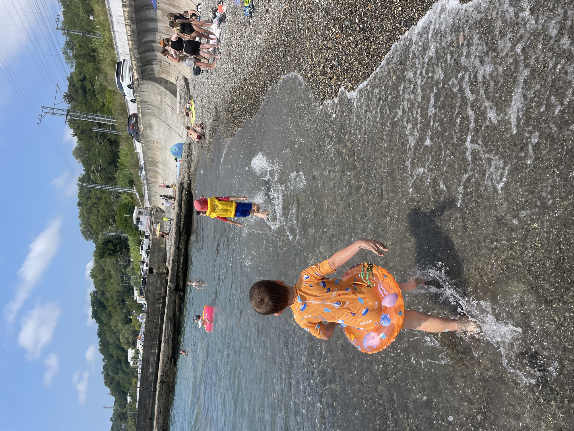 This image depicts a sunny day at a pebbled beach in Sochi, Russia. Two young boys are the primary subjects, both wading in the shallow, clear water. The boy in the foreground, facing away from the camera, wears an orange swimsuit with a playful print and a fish-themed inflatable ring around his waist. He is kicking up splashes as he walks. In the mid-ground, a second boy, also facing away, wears a bright yellow life vest and blue shorts. He is further into the water, with more significant splashes around him, suggesting he is running.
Other people are visible in the background on the beach and in the water. A group of women in swimwear are gathered on the shore, some standing and others sitting or lying on towels. Further out in the water, several other individuals, including adults and possibly children, are present. The beach is lined with a concrete embankment, behind which are trees and a road with parked cars. Overhead, power lines and poles are visible against a blue sky with scattered white clouds. The overall scene is one of recreational activity on a warm day at the seaside.