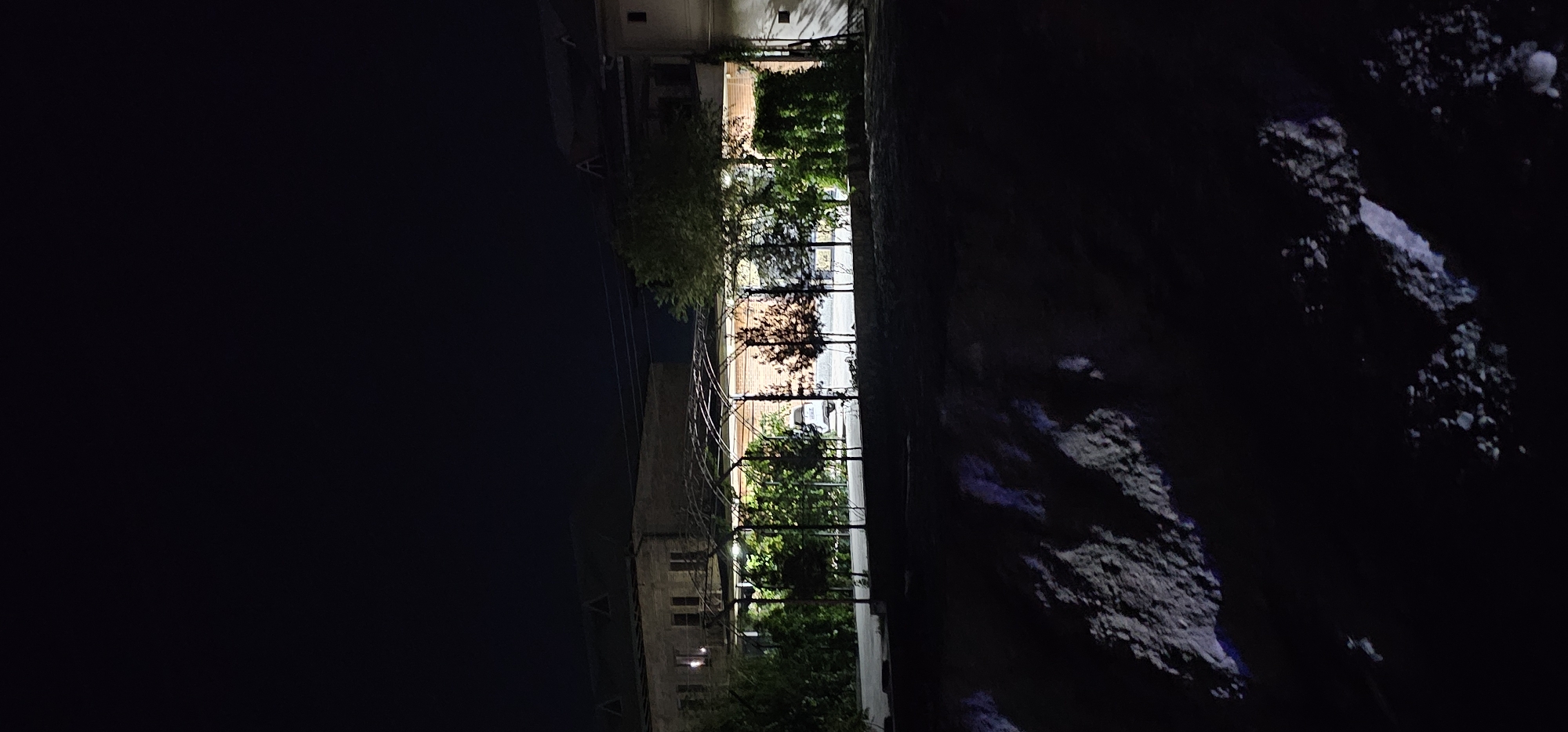 In Sheyhancha, Uzbekistan, an outdoor scene at night reveals a multi-story building with a pitched roof and multiple windows illuminated from within. In front of this structure, a covered walkway with an arched metal frame is visible, supported by pillars. This walkway shelters several trees and a white vehicle parked underneath. The area is illuminated by artificial lights, casting shadows and highlighting the vegetation. The foreground consists of a dark, rocky terrain.