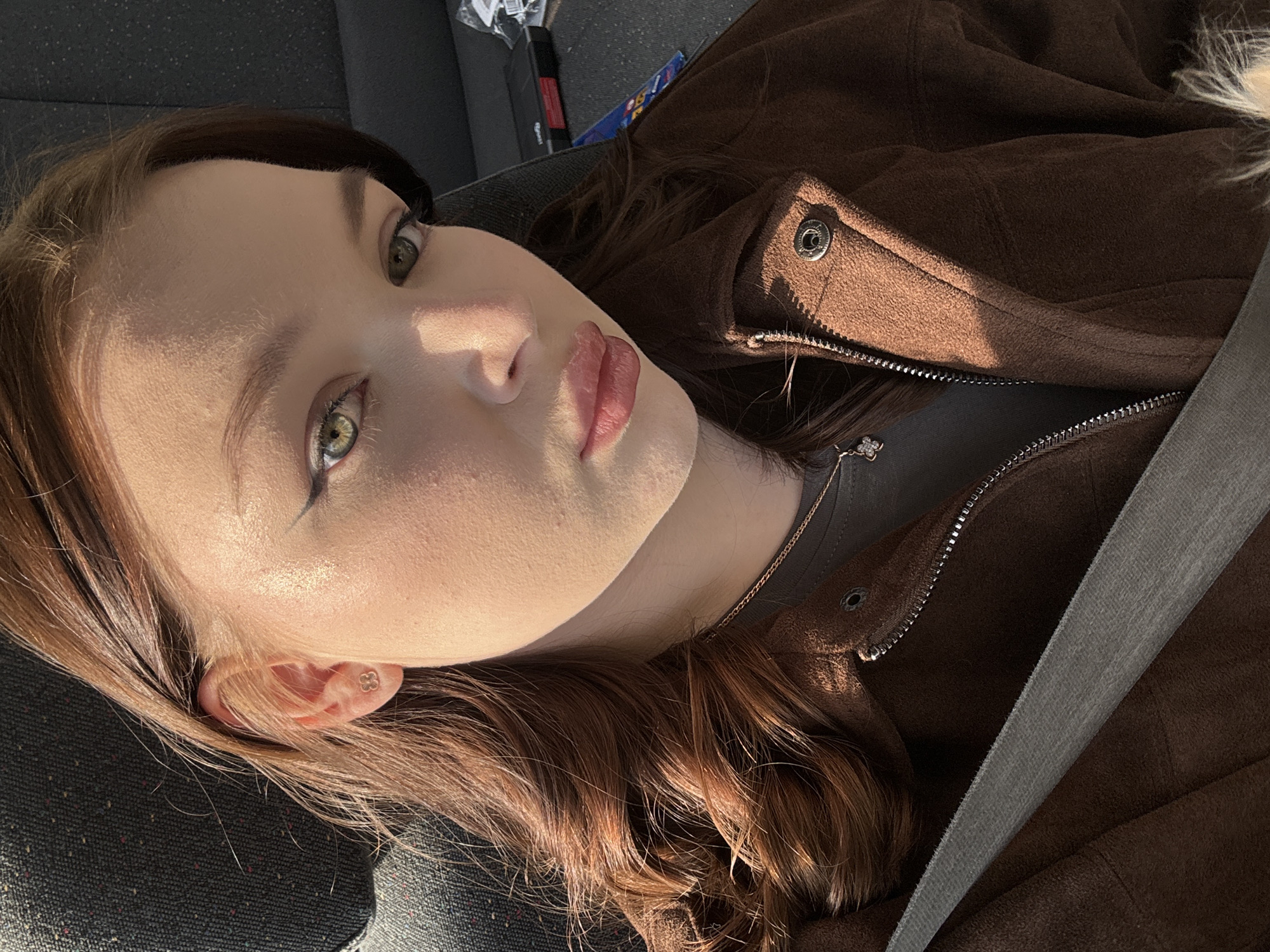 The image shows a young woman indoors, likely in a car, as indicated by the seatbelt and interior. The woman has fair skin, brown hair, and green eyes. She is wearing a brown jacket over a gray shirt and a delicate necklace. Her hair is styled with loose waves. She has a subtle eyeliner and lip color. Light is shining on her face. The background includes part of a car seat. There is not enough information to determine a location in Yekaterinburg, Russia.