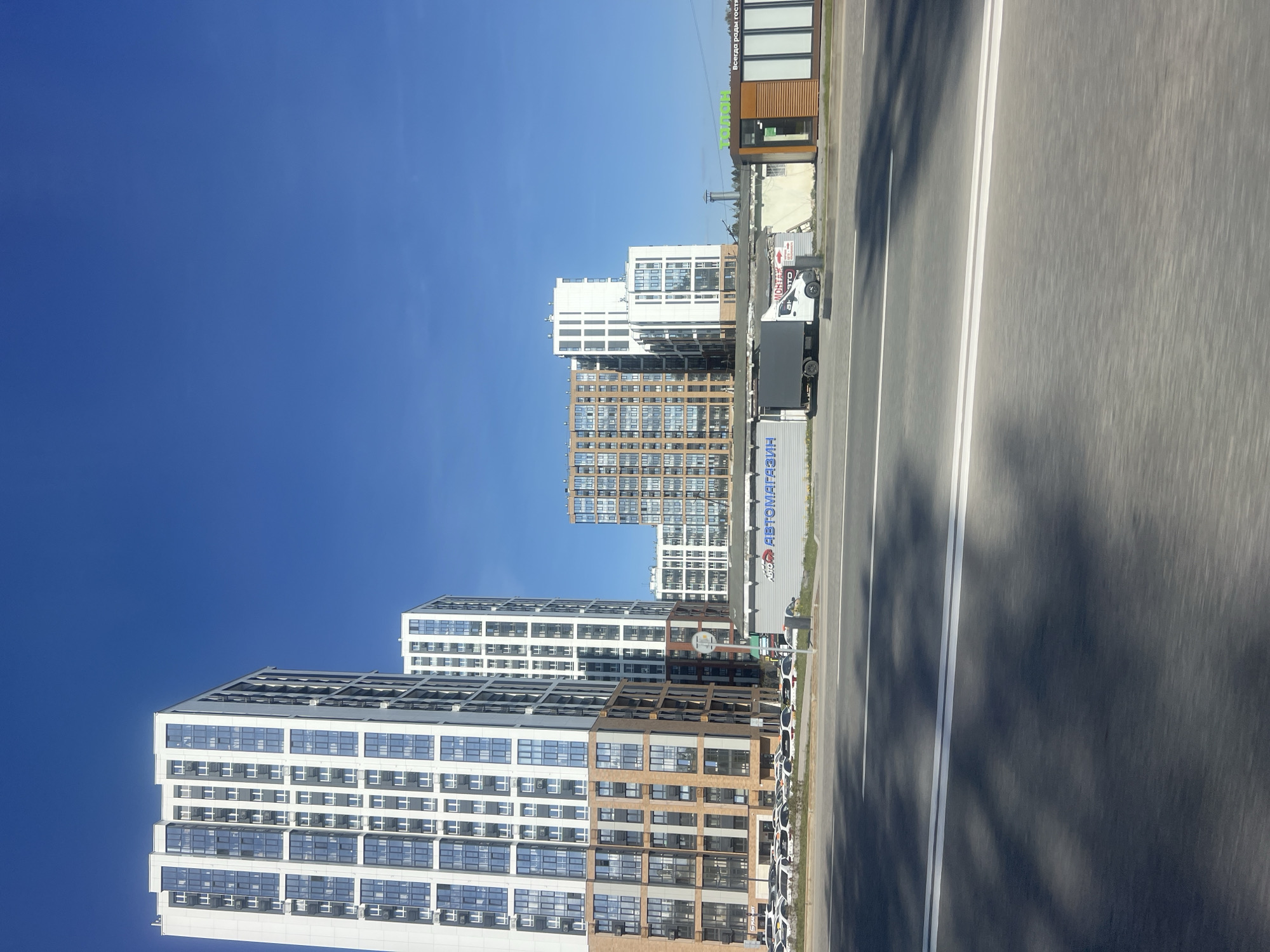 The image shows a view of a city street with modern residential buildings. The sky is clear and blue. The buildings are tall, with white and brown facades and numerous windows. A truck is parked near the buildings, partially obscuring them. A multi-lane road with white lane markings is in the foreground. The perspective suggests the photo was taken from a moving vehicle. The location is Perm, Russia.