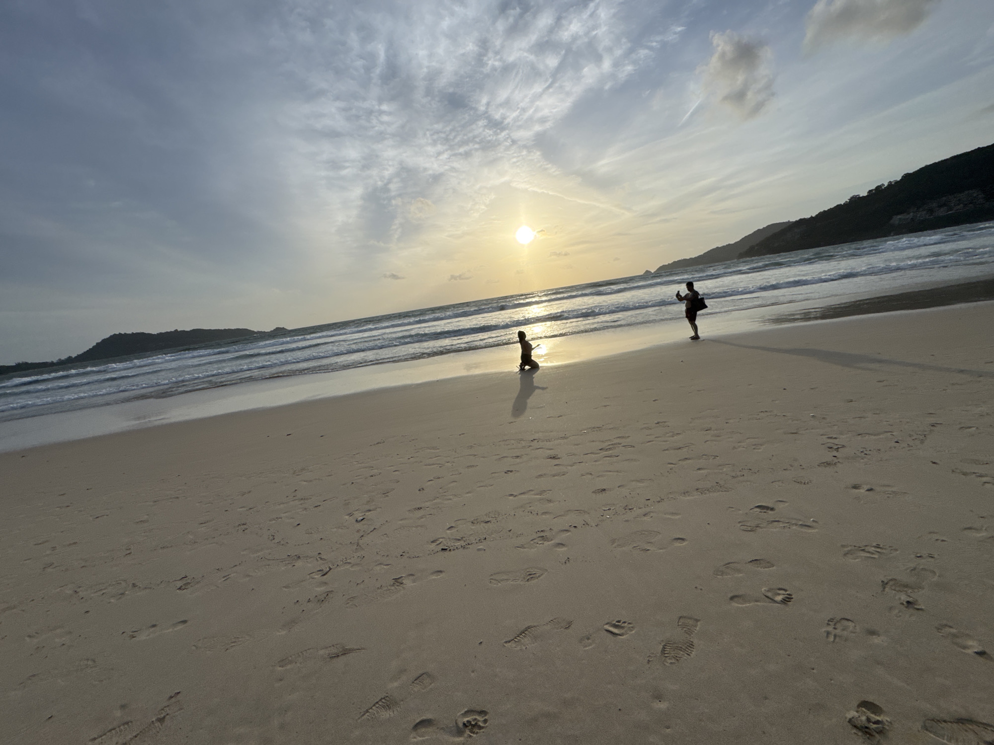 The image depicts a beach scene at sunset in Patong, Thailand. Two individuals are present on the sand. One person, dressed in dark clothing, is kneeling near the water's edge and appears to be looking out at the ocean. The second person, also in dark clothing, stands further up the beach, holding what appears to be a phone and taking a photo or video. The sun is low on the horizon, casting a warm glow across the sky and reflecting on the wet sand and ocean. Waves are gently rolling onto the shore, and the sky is filled with scattered clouds. The beach is marked with numerous footprints. In the background, hills covered in vegetation form the coastline.