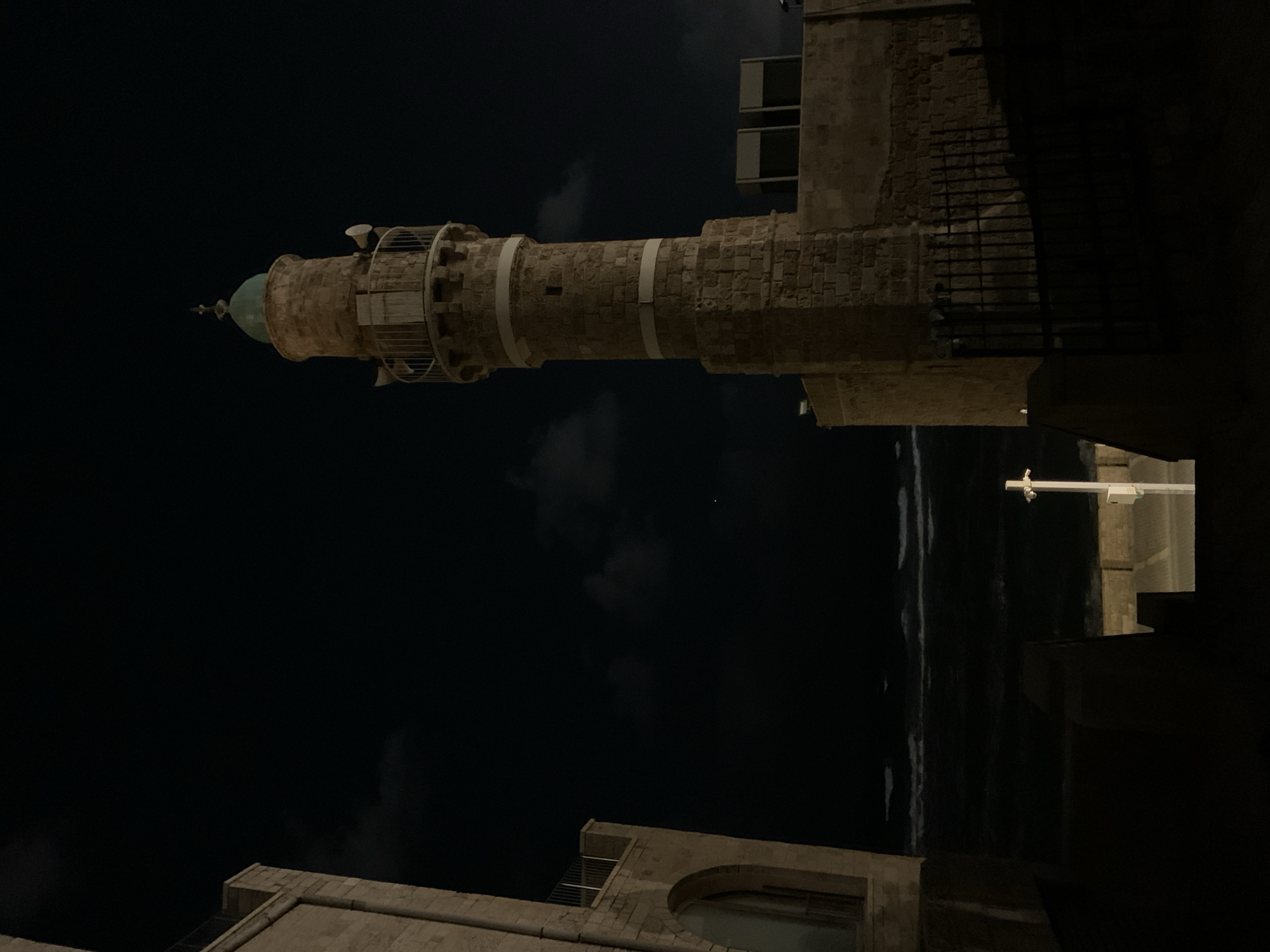 The image shows the Minaret of the Mahmoudiya Mosque in Jaffa, Tel-Aviv, Israel, at night. The minaret is made of light-colored stone. The mosque and surrounding buildings are only partially visible due to the darkness. The sea is visible in the distance, and there are some clouds in the sky.
