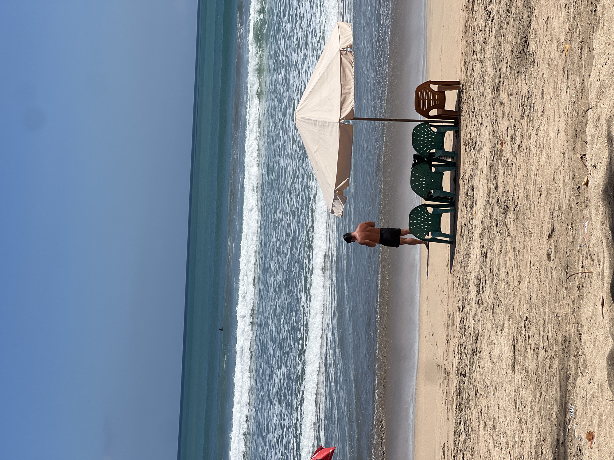 In Kuta, Indonesia, a man stands on a sandy beach near the water's edge, wearing black swim trunks. He is facing away from the camera and appears to be holding a mobile phone. Three green plastic chairs and one brown plastic chair are arranged under a beige beach umbrella. The ocean stretches out to the horizon with gentle waves breaking on the shore. The sky is a clear blue. A red triangular flag is visible on the left side of the frame.