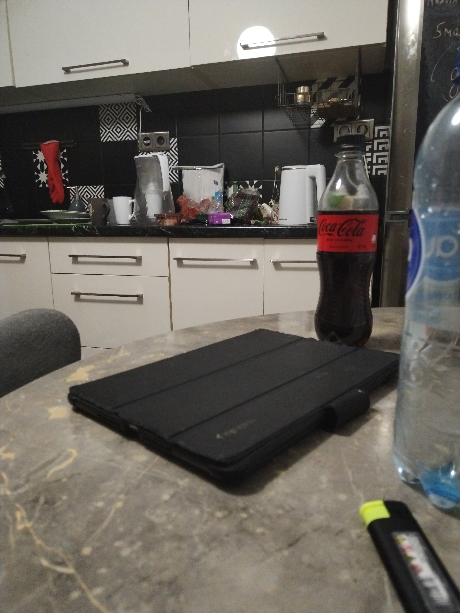 This image depicts an indoor kitchen scene in Бараўлянскі сельскі Савет, Belarus. The background features white upper and lower kitchen cabinets equipped with dark horizontal bar handles. The backsplash consists of black tiles adorned with white geometric patterns, specifically squares and star-like designs. A dark countertop is visible beneath the cabinets.
On the countertop, from left to right, a red oven mitt or glove is hanging on a rail mounted on the backsplash. Adjacent to it are a dark grey soap dispenser, a white ceramic mug, a clear pitcher, and a white electric kettle branded "Krupsser." Further right, a clear bag or container holds numerous white paper or plastic items. A small metallic shelf is suspended from an upper cabinet, holding two small metallic containers. A double electrical outlet is mounted on the black tiled backsplash. To the far right, a portion of a metallic refrigerator is visible, beside which a dark surface, possibly a chalkboard, displays faint white writing, including "5 ма."
The midground is dominated by a light grey table with subtle yellow/beige veining, extending from the bottom left to the bottom right of the frame. Part of a dark grey upholstered chair is visible on the left side of the table.
In the foreground, resting on the table, is a black tablet enclosed in a closed black case, positioned horizontally. To its right, a full 0.5L Coca-Cola bottle with a red label, displaying "Coca-Cola zero sugar" in Cyrillic, stands upright. A blurred, clear plastic water bottle is partially visible on the far right. In the bottom right corner, a black lighter with a bright yellow cap is partially visible.