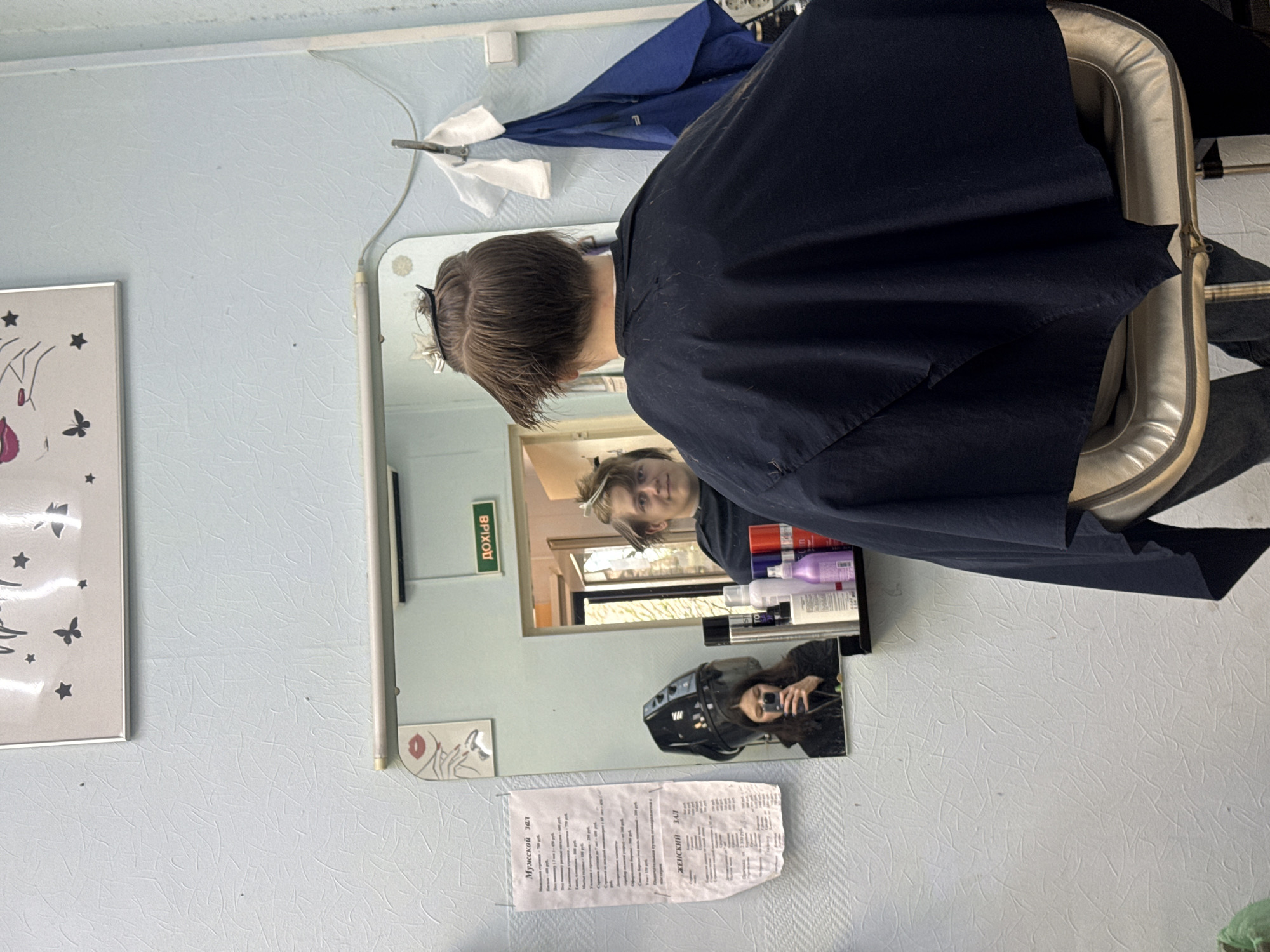 A medium shot shows a person in a hairdresser's chair with a dark cape on, seen from the back and side, with the person's face reflected in a mirror. The location appears to be a hair salon in the city of Unknown, Russia, based on the text on the paper attached to the wall below the mirror. The room has light blue walls. The mirror reflects the person's face, a shelf with various hair products, and another person holding a phone and taking a picture in a hair dryer chair. A framed picture with butterfly and star designs hangs on the wall to the left of the mirror.