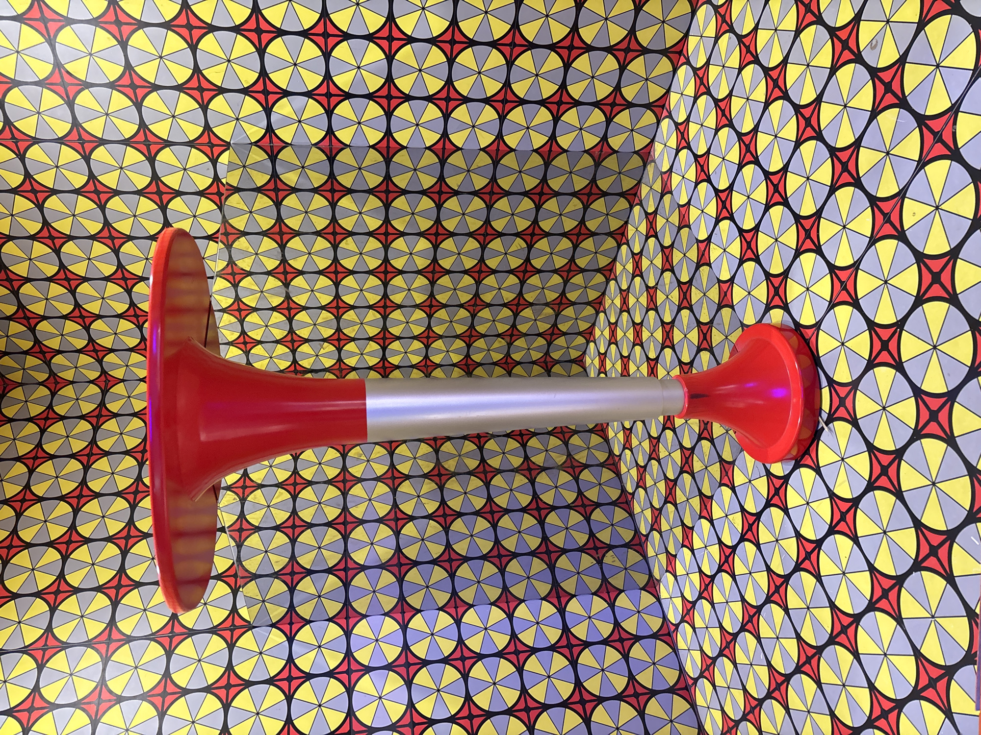 A red and silver air horn lies horizontally within a corner of a room. The corner and the walls are covered with an optical illusion pattern of yellow, gray, and red circles and stars. There is no indication in the image that it is in Jiangjin, China.