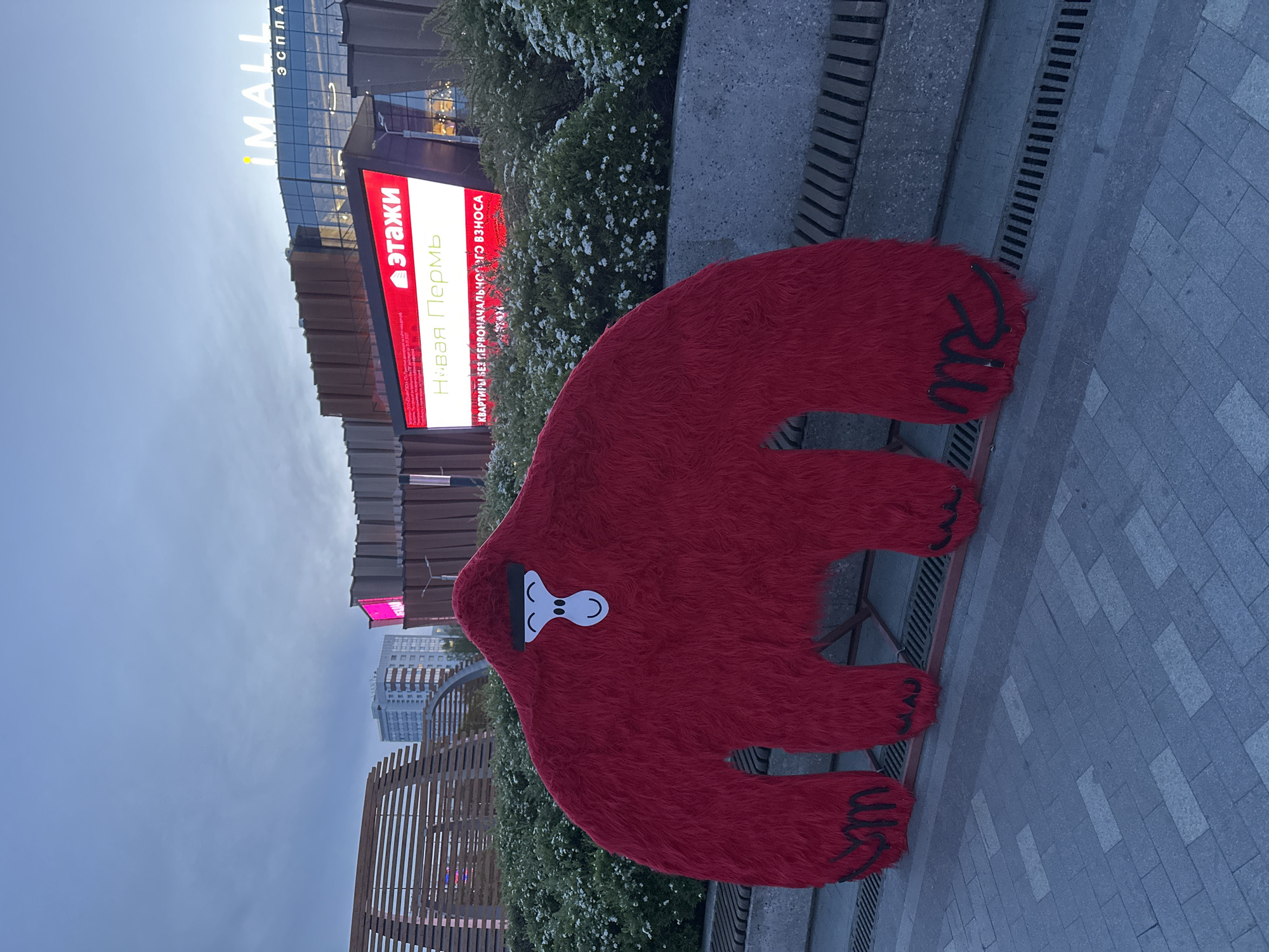 A red, furry bear-shaped sculpture stands on a paved walkway in Perm, Russia. The sculpture has a simple white and black facial design. Behind the sculpture is a low wall with shrubbery and a building with a red and white sign that reads "ЭТАЖИ Новая Пермь КВАРТИРЫ БЕЗ ПЕРВОНАЧАЛЬНОГО ВЗНОСА!". The top of the building has signage that reads "MALL". The sky is overcast.
