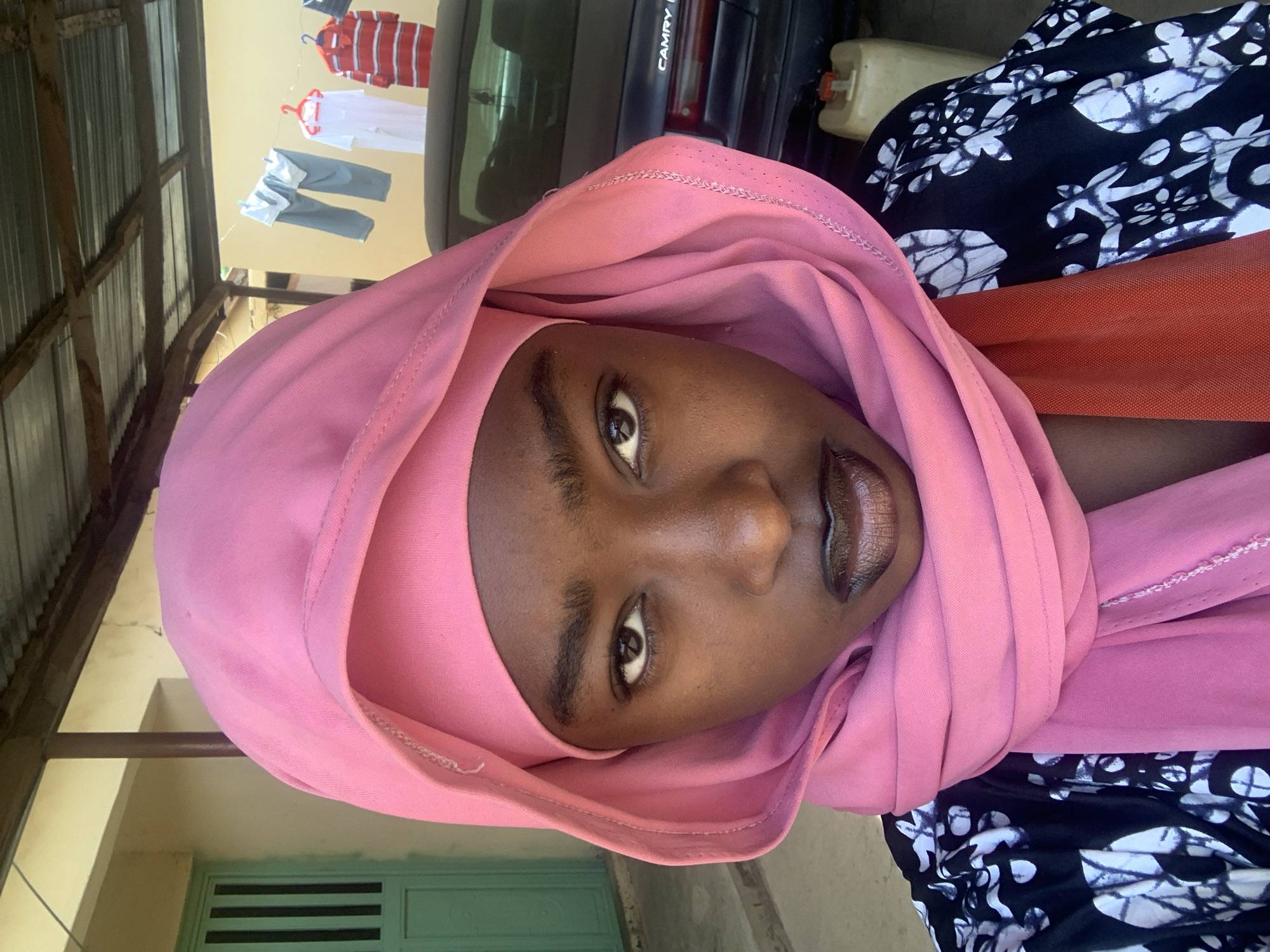A portrait of a person wearing a pink headscarf. The person has dark skin, brown eyes, and dark, glossy lips. The person is wearing a dress or top with a black and white floral pattern. In the background, there is a car, clothes hanging on a clothesline, a building with a green door, and a corrugated metal roof. The location is Rafin Par, Nigeria.