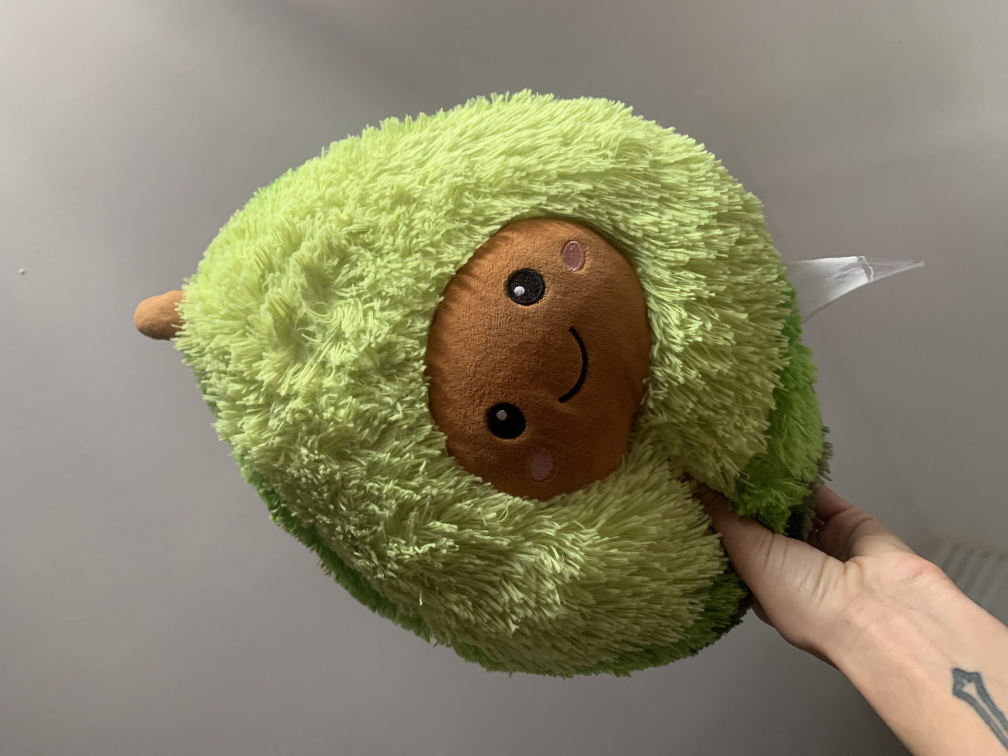 A human hand is holding a cheerful, avocado-shaped plush toy against a plain, light grey background. The plush toy is predominantly light green, covered in shaggy, soft material resembling an avocado's skin. A prominent round brown section in the center of the toy forms its face, depicted with embroidered black eyes, small pink blush marks on the cheeks, and a wide, happy black smile. A small, brown, cylindrical stem-like detail protrudes from the top left of the plush toy. A white, rectangular tag is partially visible extending from the top right side of the toy. The hand holding the toy has visible veins and subtle skin texture. A dark, stylized tattoo, appearing to be an arrow or similar symbol, is visible on the inner forearm/wrist of the hand. The lighting is soft and even, indicating an indoor setting. The scene conveys a sense of playfulness and simplicity. The location is London, United Kingdom.