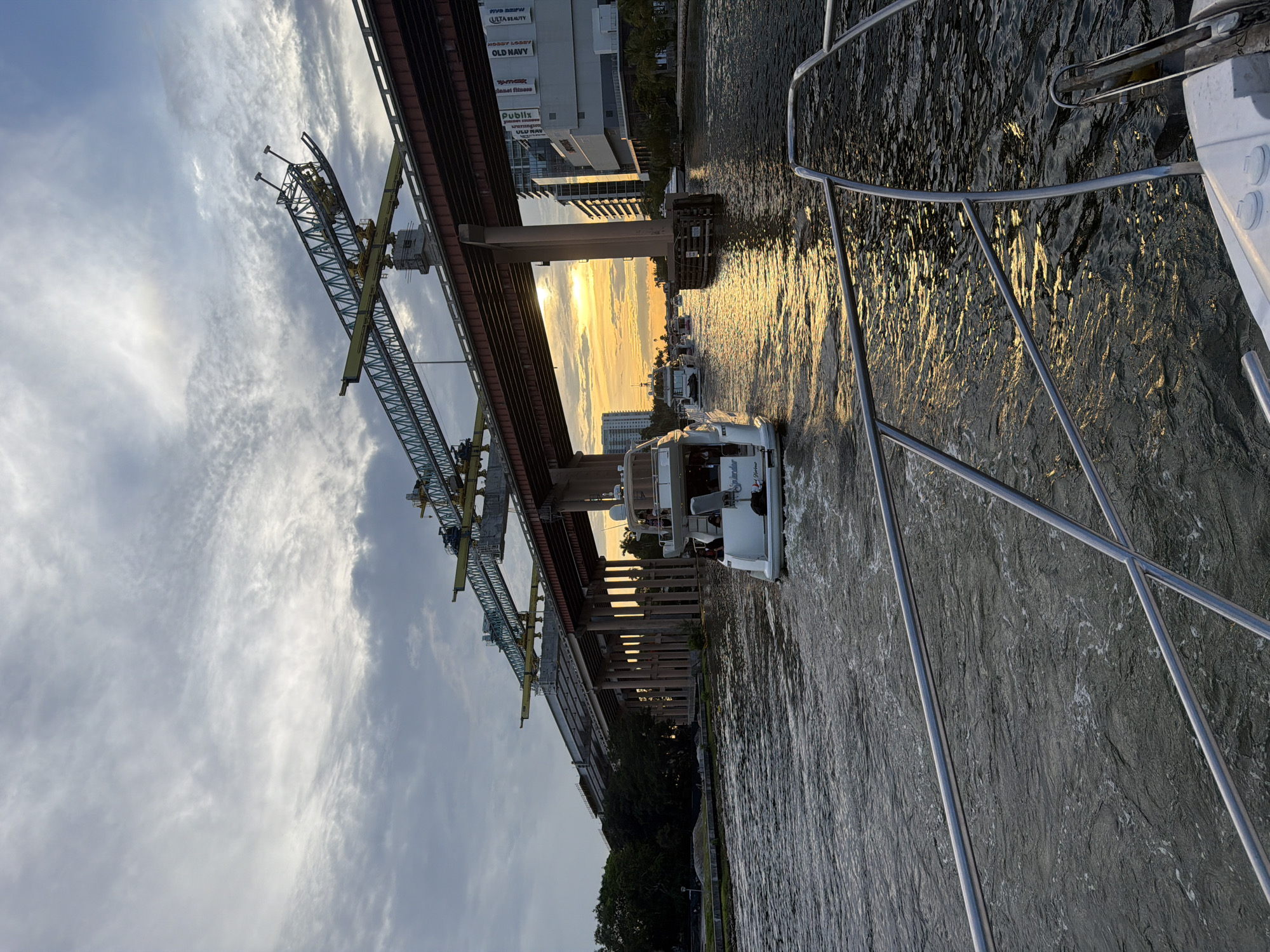 This image captures a scenic view of the New River in Fort Lauderdale, Florida, USA, during a sunset. In the foreground, the bow of a white boat with chrome railings is visible, suggesting the perspective of someone on another vessel. The river itself is a deep, dark blue, reflecting the golden hues of the setting sun and creating a shimmering effect on its surface.
Further into the scene, a fleet of boats navigates the waterway. The most prominent is a white yacht with multiple decks and people visible on board. Behind it, several other boats of varying sizes are also in motion. The river is flanked by lush green trees and vegetation on the left, and a concrete structure supporting a large overpass on the right. This overpass appears to be under construction, with visible cranes and scaffolding. The setting sun casts a warm glow through the gaps in the overpass, illuminating the water and creating long shadows. In the background, modern buildings rise above the trees, indicating an urban setting. The sky is a mix of soft grey and white clouds, with streaks of orange and yellow from the sunset. The overall atmosphere is one of peaceful transit on the water as day transitions into evening.