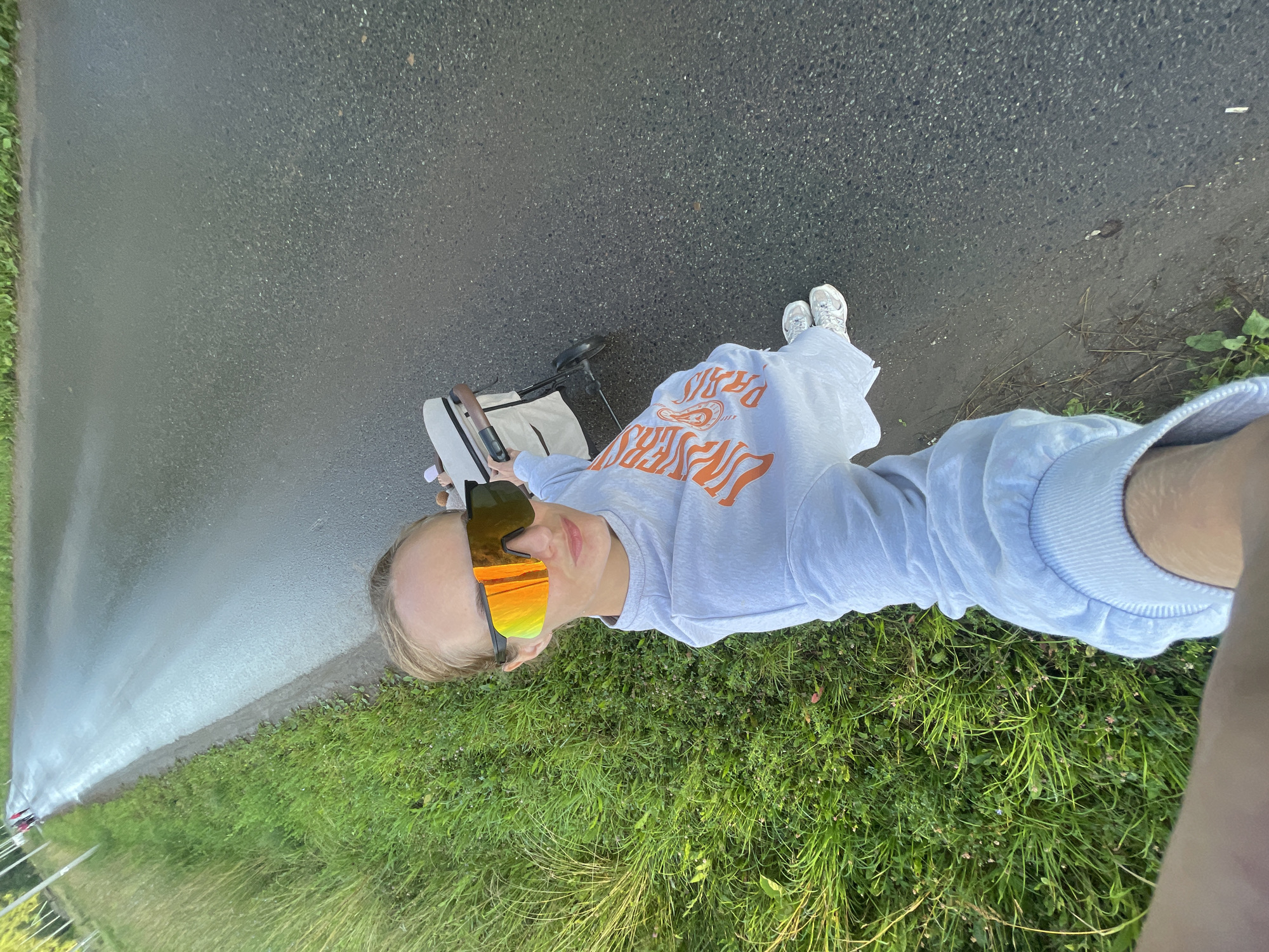 A person is standing on a wet asphalt road next to a patch of grass, holding the handle of a baby stroller. They are wearing a light blue sweatshirt with orange text that appears to say "PARIS" in a stylized font and reflective, orange and yellow sunglasses. Their feet are clad in white sneakers. The scene is outdoors, with green grass lining the road, and the road surface suggests recent rain or dampness. The lighting indicates it is daytime. The orientation of the image is such that the person appears to be lying down, with the camera held above them. A baby stroller is positioned in front of them, facing away from the camera.