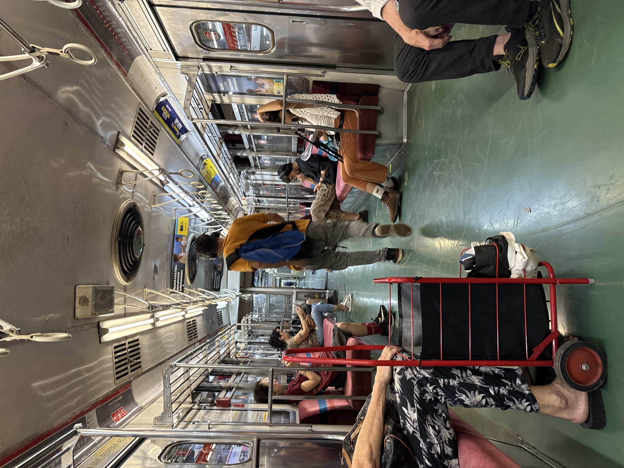 A rotated indoor shot within a subway car. Several passengers are seated, some using mobile devices. A standing man is mid-stride. A red luggage carrier, laden with clothing, stands near a seated passenger. The subway car interior features silver paneling, overhead hand grips, and advertising. The location is Buenos Aires, Argentina, deduced from the "BA" logo.