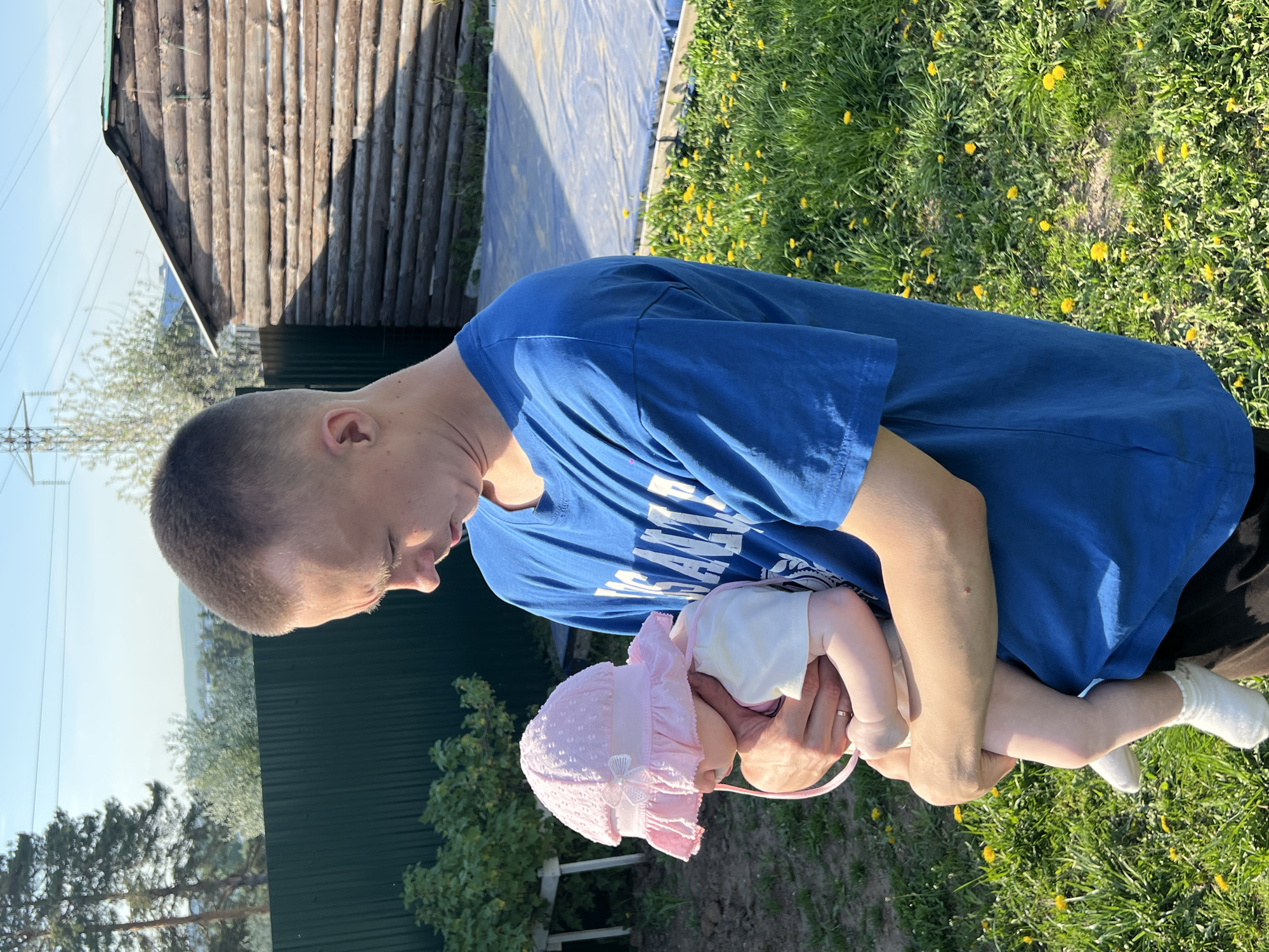 A man is holding a baby in a grassy area near some buildings. The man has short hair and wears a blue t-shirt. The baby wears a pink bonnet and white socks. The location is Chaikovsky, Russia. The grassy area has yellow flowers, and the buildings include a log cabin-like structure and a green metal fence. The photo is taken outdoors in daylight.