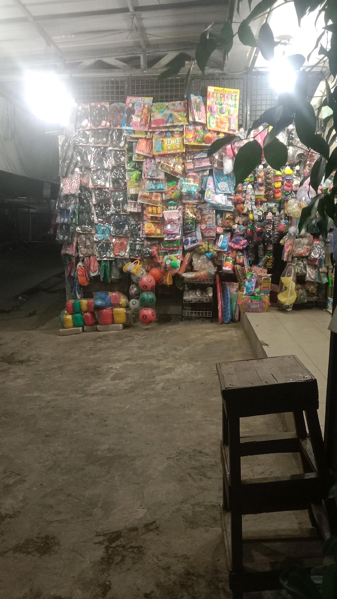 The image captures a night scene outside a small shop, possibly a market stall, in Sumberjaya, Indonesia. The shop is filled with an assortment of merchandise, primarily toys and various plastic household items. In the foreground, a weathered wooden stool or small step ladder sits on a concrete ground. Behind it, the shop's interior is visible, displaying shelves and racks densely packed with colorful packaged toys, including dolls, toy kitchens, and various other playthings. Below the shelves, colorful plastic containers and balls are stacked on the ground. A bright light source, likely from within the shop or a street light, illuminates the scene, casting shadows and creating a somewhat dim and atmospheric environment. The overall impression is of a bustling local marketplace, perhaps one that operates into the evening.