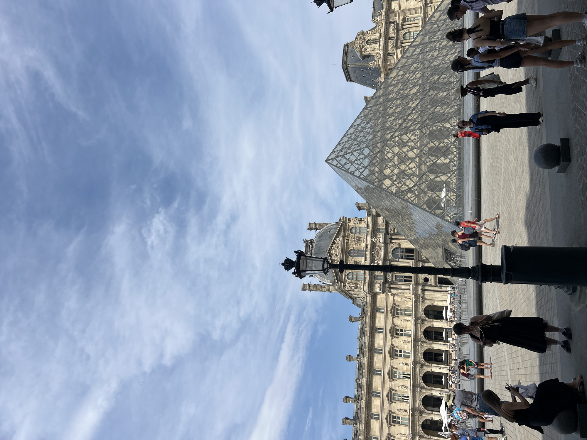 The image depicts the exterior of the Louvre Museum in Paris, France, on a partly cloudy day. The iconic glass pyramid entrance is prominently featured in the center-right of the frame, reflecting the sky and surrounding architecture. To the left of the pyramid is the historic Louvre Palace, with its classical facade, arched windows, and ornate decorations. A traditional black lamppost stands in the foreground, slightly to the left of the center.
Several people are scattered across the plaza in front of the museum. They appear to be tourists, some standing in groups, others walking, and a few are seated. Many are dressed in casual summer clothing. Some individuals are holding phones, likely taking pictures or navigating. The ground is paved with stone, and there is a decorative water feature visible behind the pyramid. The sky is a vibrant blue with scattered white clouds. An airplane is visible as a small white speck in the upper left portion of the sky.