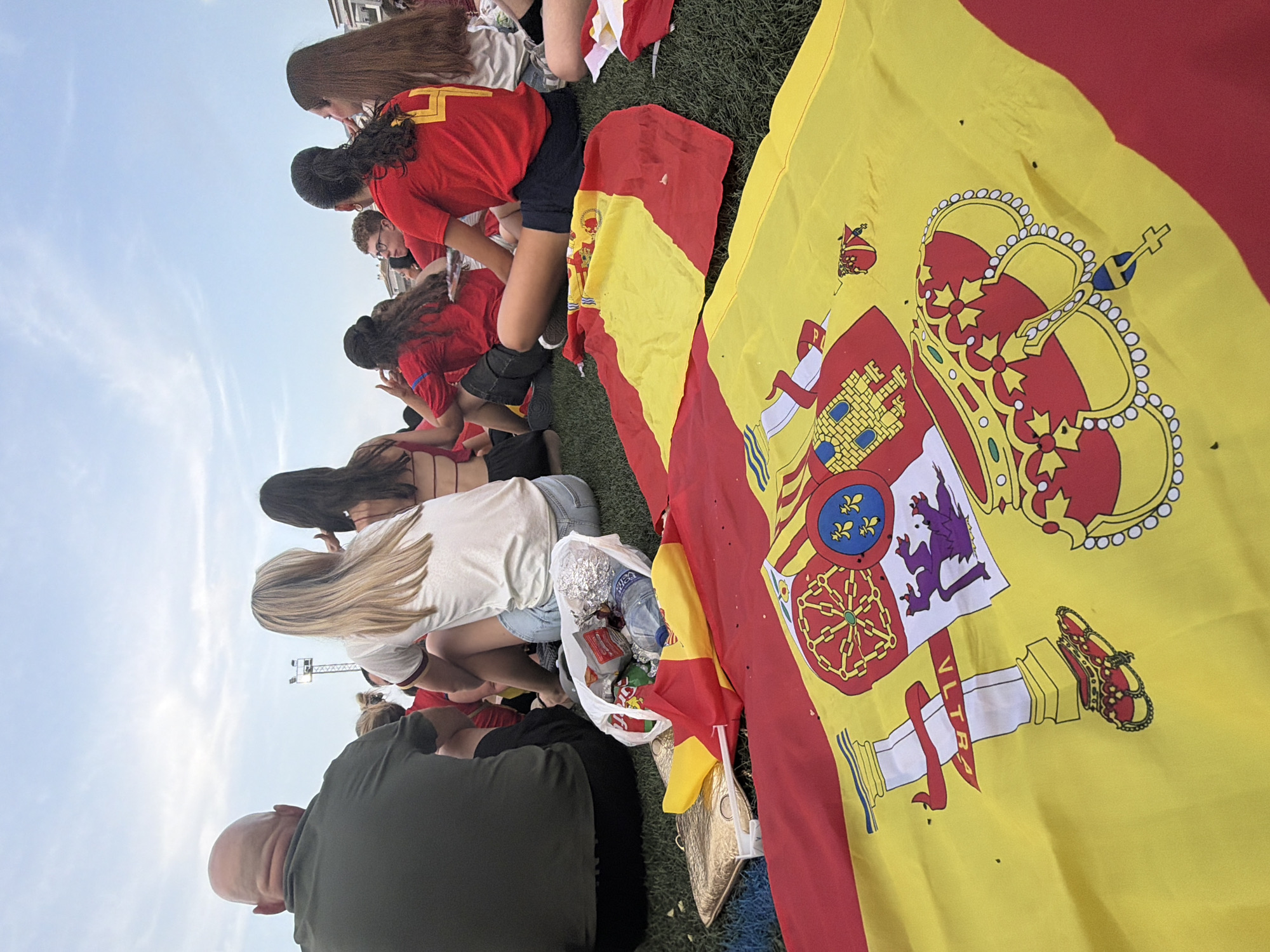The image shows a group of people sitting on a grassy field, with Spanish flags in the foreground. The people are facing away from the camera, and most of them are wearing red shirts. The Spanish flag is prominently displayed, with its red and yellow stripes and coat of arms. The sky is visible in the background, and it appears to be a bright, sunny day. The overall scene suggests a gathering of fans or supporters, possibly watching an event.
Based on the visual evidence provided, the image can be localized to the city of Dénia, Spain. Specifically, the flags and the red shirts worn by many individuals suggest a connection to Spanish national pride, possibly related to a sporting event or a national celebration. The grassy field and open sky indicate an outdoor public space commonly used for such gatherings. While the exact location within Dénia cannot be determined from the image alone, it is consistent with public parks or open areas in the city.