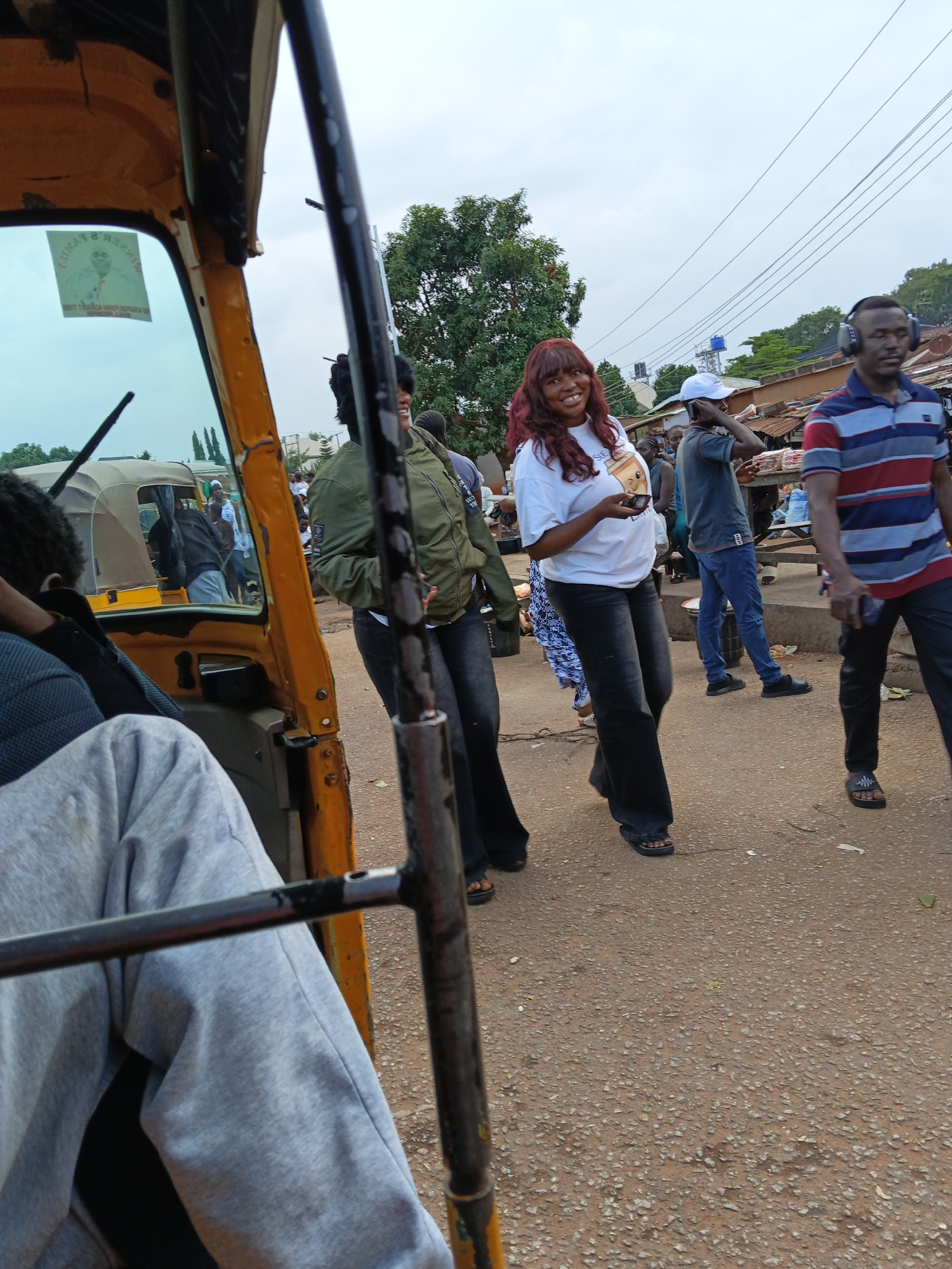 A group of people are gathered on a dusty street in Bukuru, Nigeria. In the foreground, a person sits inside a yellow auto-rickshaw, their legs visible wearing grey sweatpants. A black metal bar runs across the image in front of them. To the right of the auto-rickshaw, a woman with long, reddish-brown hair smiles as she holds a small object in her hand. She is wearing a white t-shirt and dark pants. Behind her, another woman in a green jacket stands and looks to her right. Further back, a man wearing headphones and a striped shirt is walking away from the camera. Other individuals are visible in the background, some standing near stalls and structures that line the street. The environment appears to be an outdoor market or busy street with signs of commercial activity. The sky is overcast, suggesting a cloudy day.