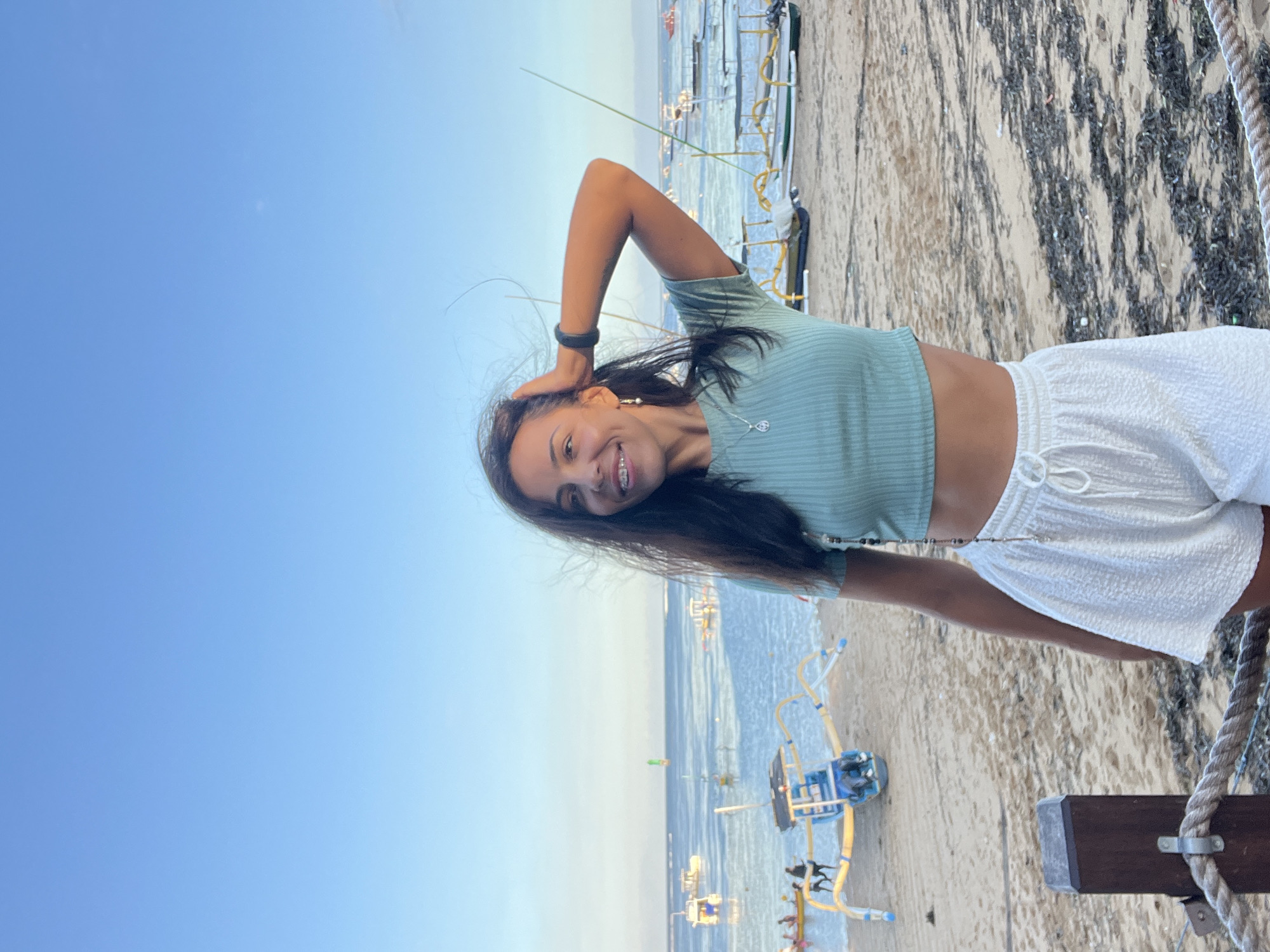 The image depicts a woman standing on a beach in Denpasar, Indonesia. She is wearing a light green short-sleeved crop top and white shorts. She is smiling, with her left hand behind her head and her right arm positioned downward. Her hair is dark and flowing. In the background, there is a clear blue sky transitioning to a pale blue horizon where the sky meets the ocean. Several boats are visible offshore, as well as beachside structures. The foreground includes a sandy beach, seaweed, and part of a roped fence with a wooden post.