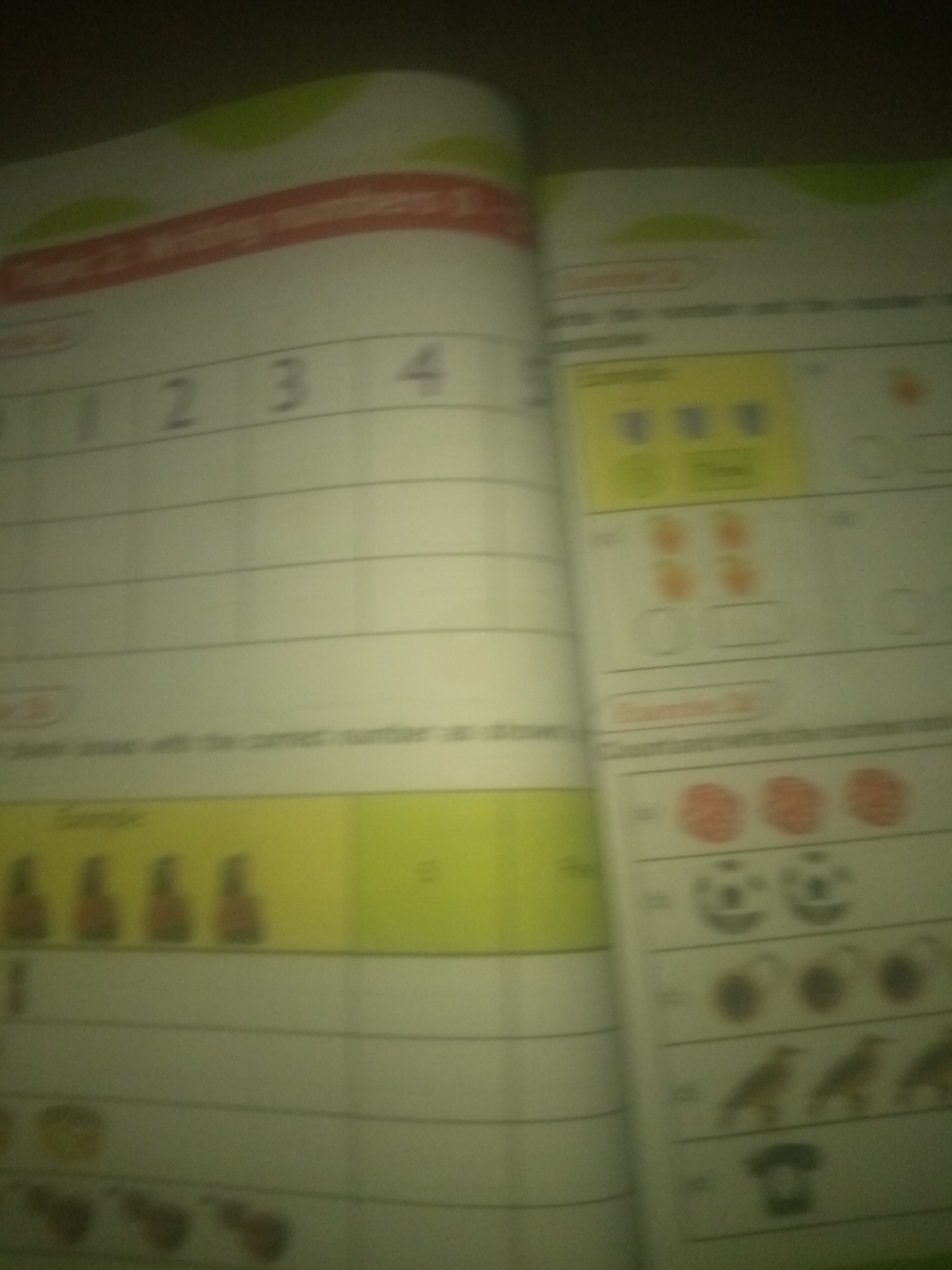 The image shows two pages of a workbook, with one page on the left and another on the right. The left page has a title at the top, followed by a grid with numbers 1 through 5 at the top of the columns. Below this, there are horizontal lines for writing. There is a section with a yellow background that contains images of bottles and a number "3".
The right page has various exercises with images and spaces for answers. There are exercises involving counting objects like apples, balls, and birds. Some exercises have boxes with numbers or empty spaces. The overall context suggests a workbook for early learning, likely focusing on counting and number recognition. The image is blurry and taken at an angle, making it difficult to read all the text and clearly identify all the objects.