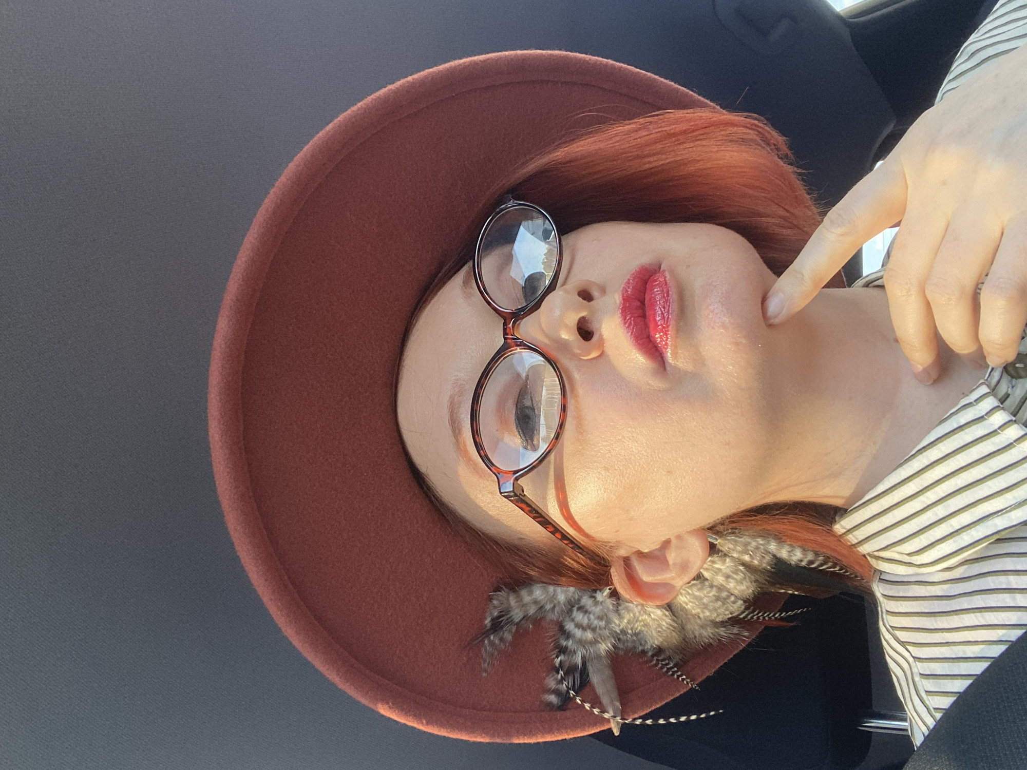 The image presents a close-up portrait of a person, rotated 90 degrees counter-clockwise, so the subject's head is facing to the right. The individual has fair skin and vibrant red or auburn hair, partially visible beneath a stylish, wide-brimmed hat in a burnt-orange or rust color. They are wearing round glasses with tortoise-shell frames, which show reflections of bright light. Their lips are painted with glossy red lipstick.
The person's right hand, with an extended index finger, gently touches their chin or jawline. A striped shirt, predominantly white or cream with thin olive-green or grey stripes, is visible around their neck and shoulder. Decorative feathers, featuring shades of grey, brown, and black patterns, are tucked into the hat near their ear.
The background is dark, out-of-focus, and appears to be an interior space, possibly a car, suggested by the subtle texture and dark tone. Bright, natural light illuminates the subject from above and slightly to the side, creating prominent highlights on their skin, lips, and the lenses of their glasses. The expression is poised and somewhat contemplative. No visible text is present in the image. The location context provided is Yekaterinburg, Russia.