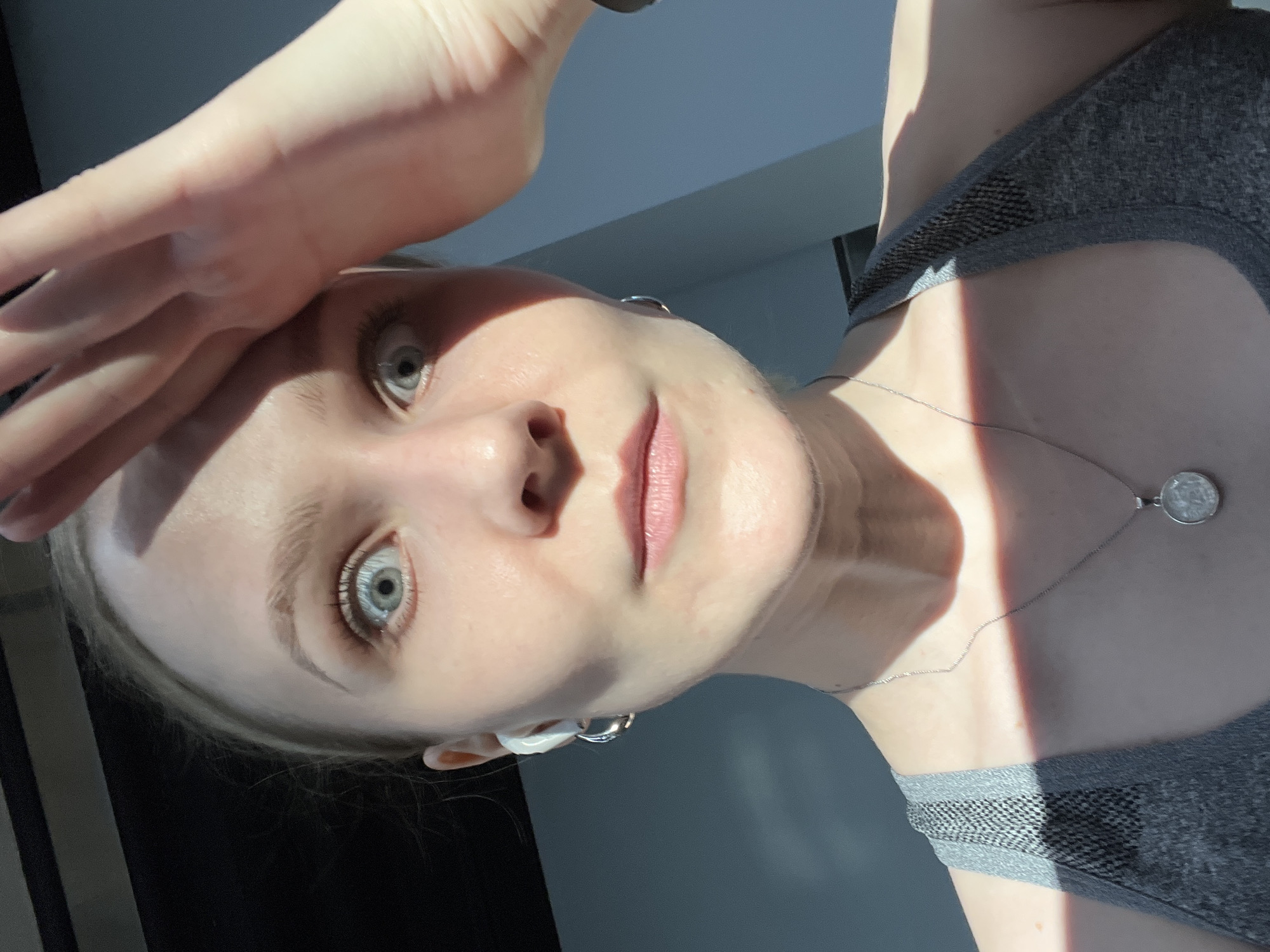 A woman with light skin and blue eyes is angled toward the camera. She is wearing a gray tank top and a silver necklace with a circular pendant. Her hand is raised to her forehead, blocking some of the sunlight. She is wearing silver hoop earrings. The location is indiscernible from the image.