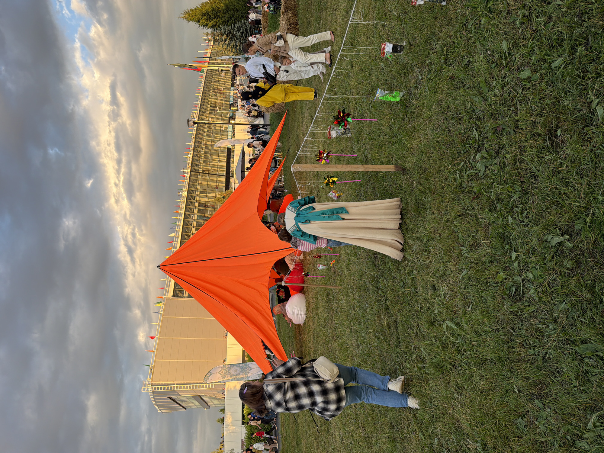 An outdoor event is taking place on a grassy field, likely in a park or fairground. In the background, a modern, multi-story building with a glass facade and numerous small flags displayed on its roof and along its exterior is visible. The sky is partly cloudy with dramatic lighting.
In the foreground, a large, bright orange tent is set up. Underneath the tent, several people are seated on large beanbag chairs. To the right of the tent, a mannequin displays a cream-colored dress with a teal sash. A string with colorful pinwheels and small hanging objects, possibly prizes or decorations, stretches across the foreground.
Several individuals are walking and standing in the scene. A woman in a black and white plaid shirt and jeans is walking away from the camera, holding a phone. A group of three people, including a woman in a yellow jumpsuit, a man in a white shirt and pants, and a young child in white, are standing to the right. Other attendees are scattered throughout the scene, with more people and structures visible further in the background. The overall atmosphere suggests a casual outdoor gathering or festival.