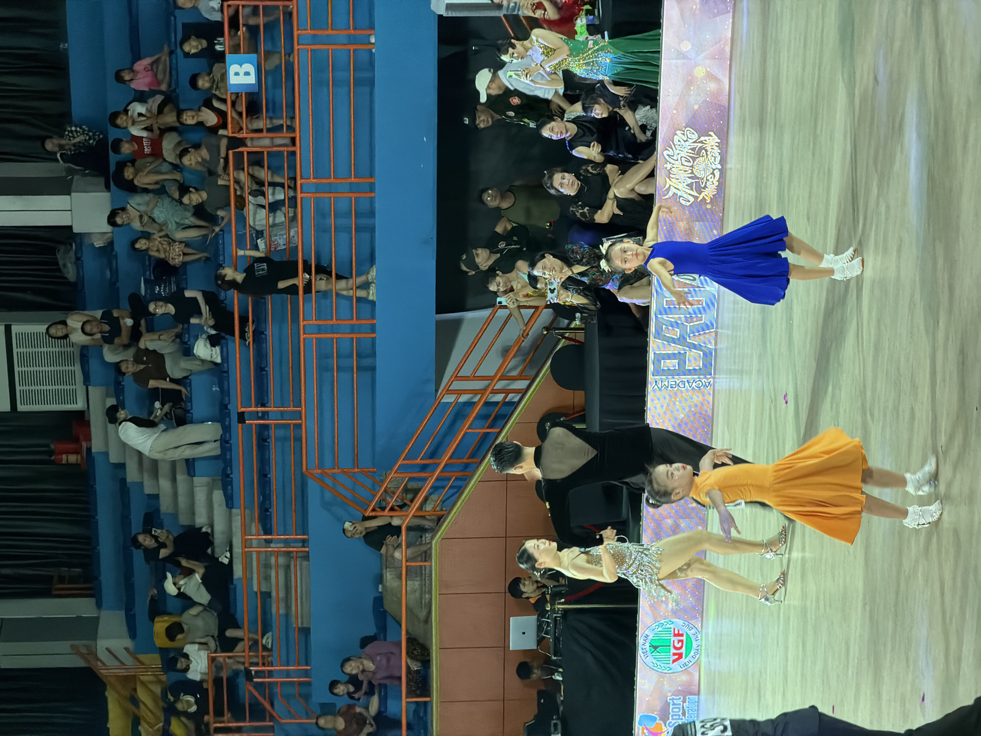 In a brightly lit indoor arena in Hanoi, Vietnam, a ballroom dancing competition is underway. The foreground features two young girls in dazzling costumes, one in a vibrant orange dress and the other in a royal blue one, mid-performance with outstretched arms and poised stances. Behind them, a couple in formal dance attire, the man in a black velvet costume and the woman in a glittering silver dress, are also engaged in their performance. Spectators are seated in tiered blue bleachers, some actively watching the dancers and others engrossed in their mobile phones. The arena itself is marked with sponsor banners and decorative elements, suggesting a professional event. The overall atmosphere appears to be one of focused energy and lively entertainment.