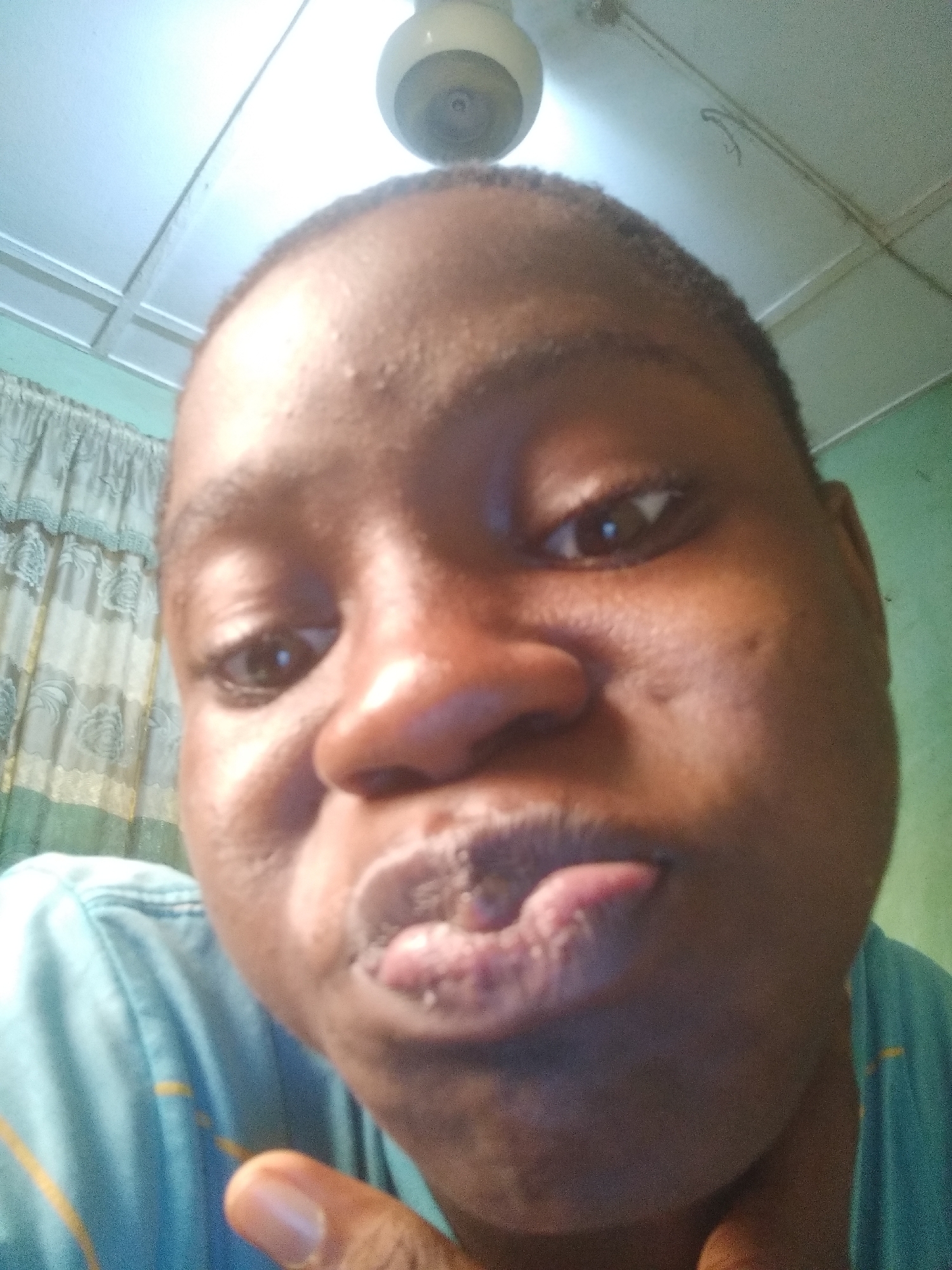 The image is a close-up selfie of a Black person with short hair, light brown eyes, a large nose, and full lips slightly pursed. The person is wearing a light blue t-shirt. The background includes a white ceiling with a ceiling fan and a curtain with a floral pattern. There is no indication of location in Shangisha, Nigeria in this image. The focus is solely on the individual's face and immediate surroundings.