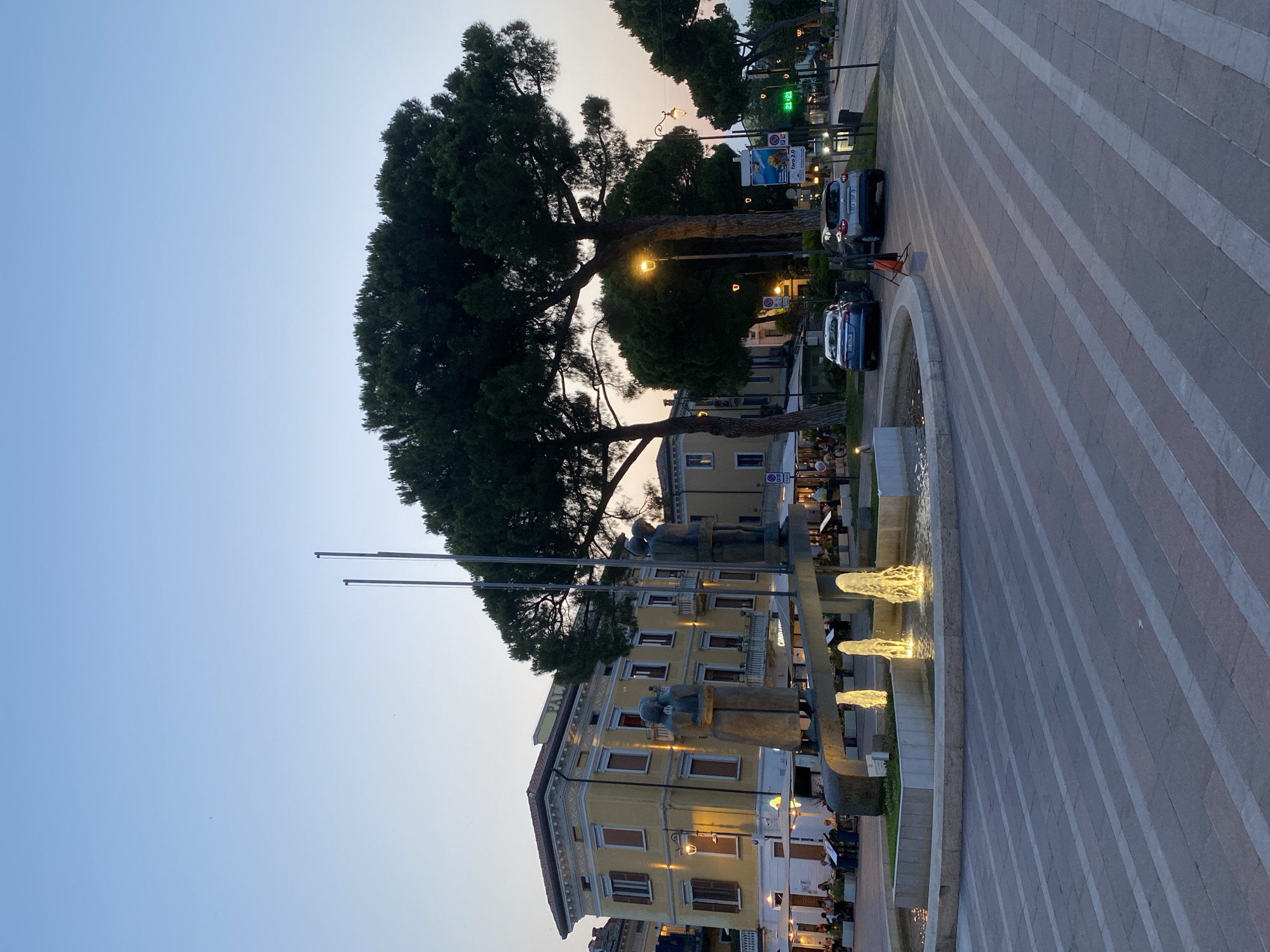 This is a daytime, low-angle, outdoor, eye-level shot of a public square in Desenzano del Garda, Italy. In the foreground, there is a modern sculpture that appears to be made of metal, with abstract figures. In front of the sculpture is a fountain with illuminated jets of water. The plaza is paved with large rectangular stones in a striped pattern. Behind the sculpture is a building with yellow facade, balconies and brown shutters. Adjacent to the building, there are several large trees. In the middle ground, two cars are parked. On the right side of the image, there are more trees, street lights, and a traffic light showing 21:21. In the background, there are more buildings and people sitting at outdoor tables. The sky is a pale blue, indicative of dusk.