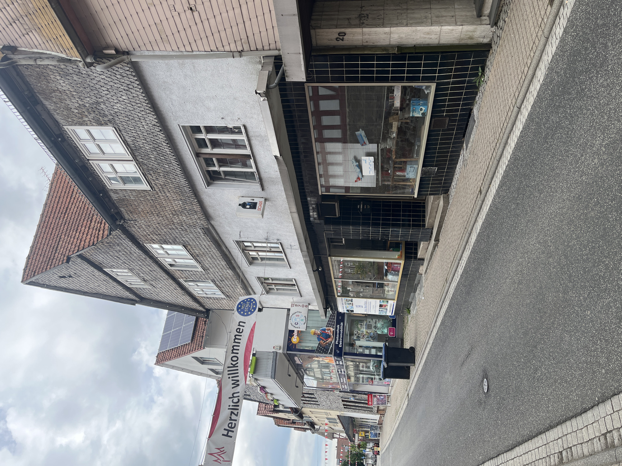The image depicts a street scene in Neustadt, Germany, under a cloudy sky. A banner that reads "Herzlich willkommen" (Welcome) is strung across the street. The scene features buildings with traditional German architecture, including steeply pitched roofs covered in wooden shingles and tile.
On the right side of the street, a building with a light gray facade has a shopfront with a black tiled exterior and large display windows. The windows display various items, including what appear to be model airplanes and boxes. The building's entrance is marked with the number "20".
To the left of this building, another shopfront is visible, featuring a blue and white facade with the text "Photovoltaik Elektromobilität" and an image of a worker in a hard hat. This shop appears to be selling solar power and electric mobility solutions. Two dark-colored trash bins are placed in front of this shop.
Further down the street on the left, more buildings and shopfronts are visible, suggesting a commercial area. A red tiled roof and solar panels are visible on one of the buildings. The street itself is paved with asphalt and bordered by a sidewalk made of cobblestones. The overall impression is of a quiet, European town street.