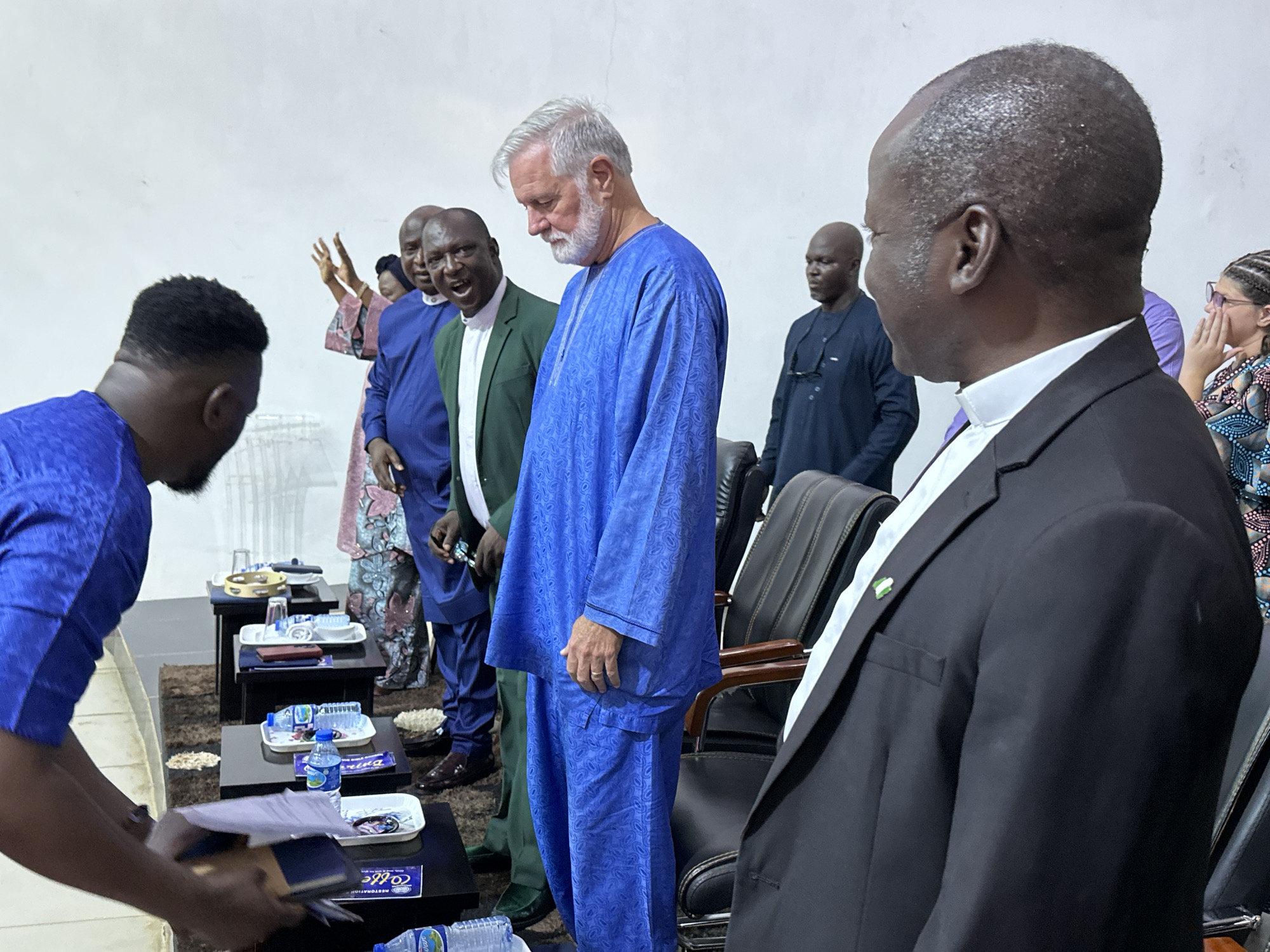 The image depicts a gathering of men and a woman in what appears to be an indoor event, possibly a religious or official function, set in Yelwa, Nigeria. The scene is focused on several individuals, predominantly men dressed in traditional attire and suits.
In the foreground, a man in a black suit with a white collar is seen from the side, his gaze directed away from the camera. To his left, another man in a vibrant blue traditional outfit with a patterned design is prominently featured, looking downwards with his left hand resting on his hip. Behind him, a man in a green suit over a white shirt is partially visible, his mouth open as if speaking. Further in the background, several other men are present, some in blue traditional wear and others in darker suits. A woman in a patterned dress and with braided hair is also visible in the far right, her hand near her face.
The environment suggests an indoor setting with a plain, light-colored wall in the background. Several small, dark tables are arranged, holding items such as bottled water, small white trays with what appear to be snacks or offerings, and perhaps some documents or booklets. The flooring is covered by a rug. The overall impression is of a formal or semi-formal meeting or ceremony.