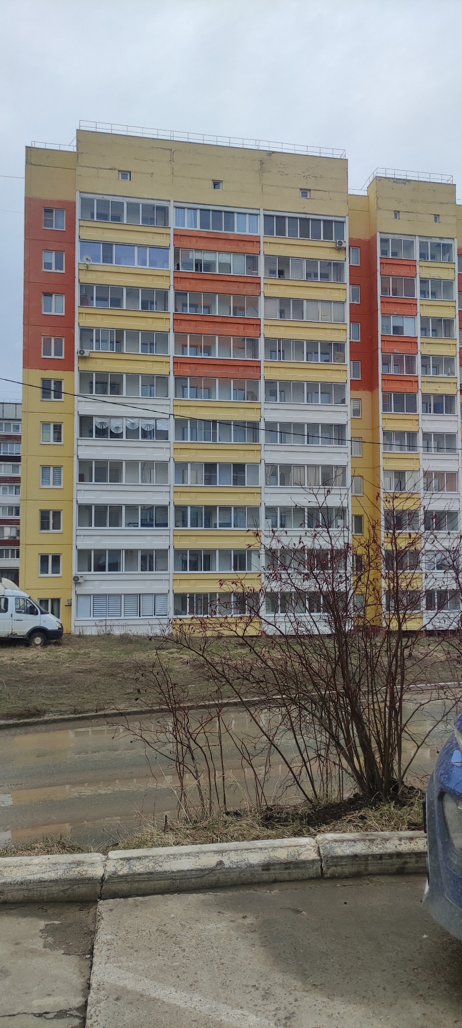 The image shows a multi-story apartment building in Chaikovsky, Russia. The building has a predominantly yellow facade with sections painted orange and white. The windows are rectangular, and there is a railing on the roof. In front of the building, there is a patch of grass and bare bushes. The foreground shows a concrete curb and a section of a wet street or pavement. A white van is parked on the left side of the building, and the front of a blue car is visible on the right. The sky is overcast.