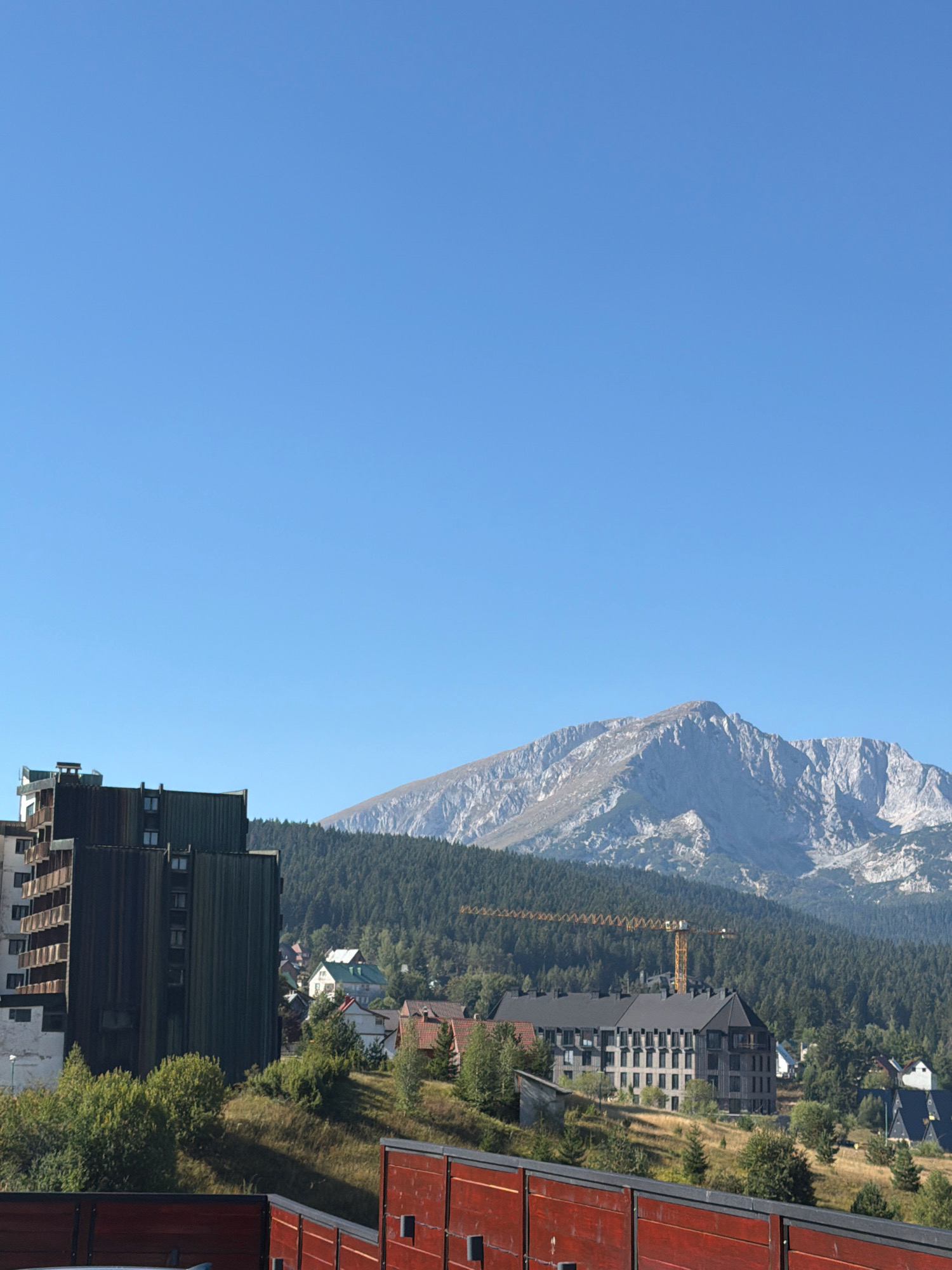 The image captures a panoramic view of the city of Žabljak, Montenegro, dominated by the imposing Durmitor mountain range in the background. In the foreground, a dark, multi-story building with balconies and a light-colored adjacent structure stands prominently. To the right of this building, nestled amongst a dense forest of coniferous trees, are several smaller residential buildings with red and green roofs. Further into the scene, a large, modern, dark-colored building with numerous windows is visible, and a yellow construction crane is situated within the treeline. The sky above is a clear, bright blue. A red wooden fence runs along the bottom of the image, obscuring the lower portion of the scene. The overall impression is of a mountainous town under a clear sky, with signs of ongoing construction.