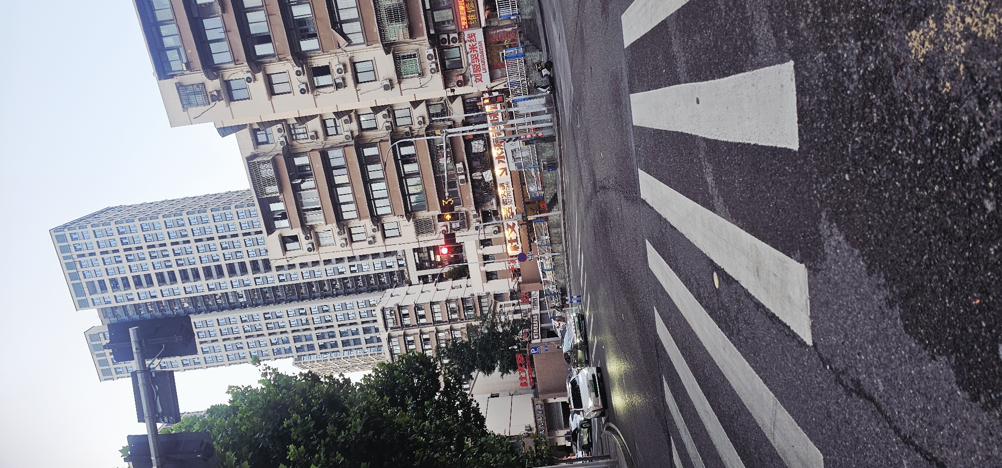 The image depicts a street scene in Yuzhong District, Chongqing, China. Several buildings of varying heights, with many windows and air conditioning units, line the street. Street-level businesses are indicated by vertical signage in Chinese characters. A red traffic light is visible. The foreground shows a crosswalk with white stripes painted on the asphalt road surface. A row of cars is parked along the street. Trees with green foliage partially obscure the view. The sky is overcast.