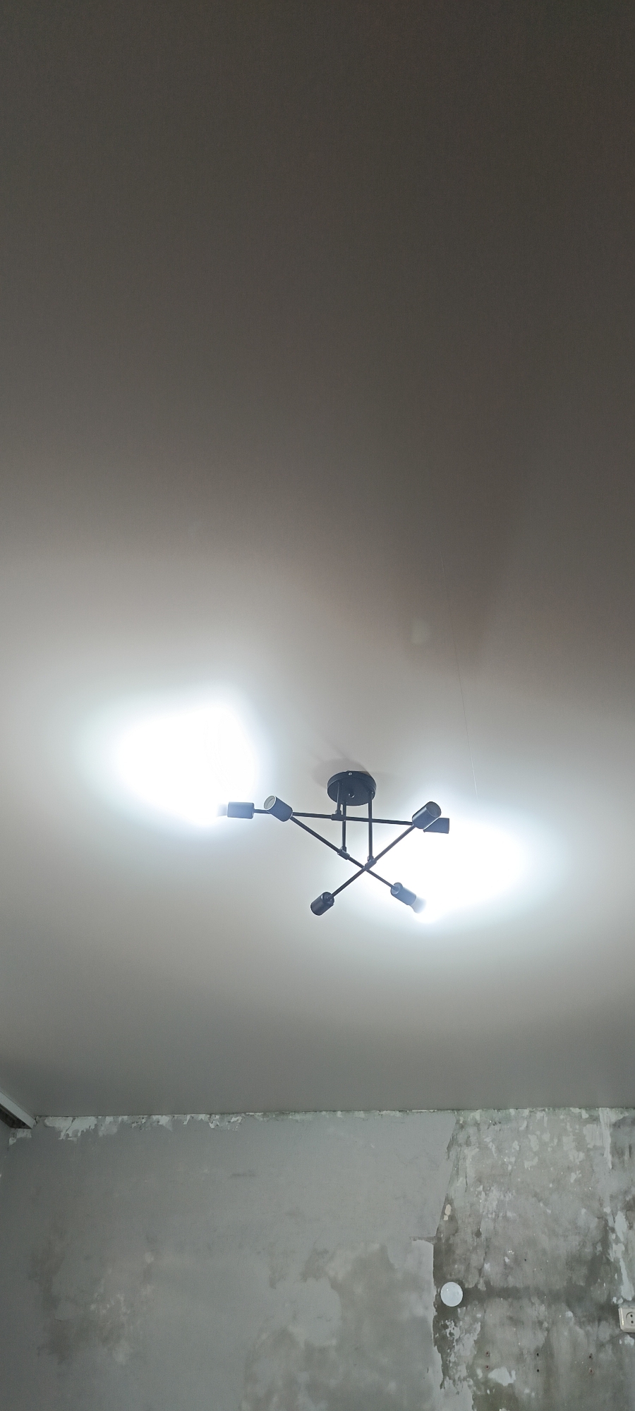 The image shows a modern, black spider-like chandelier hanging from a white ceiling. The chandelier has multiple arms extending outwards, each ending in a light fixture. The lights are on and casting a bright glow on the ceiling. Below the ceiling, a wall is visible, painted in a muted grey color. The wall appears to be unfinished or in disrepair, with patches of peeling paint and textured surfaces. There is a white circular fixture mounted on the wall, possibly an electrical outlet or a light switch. The overall scene suggests an indoor setting, likely a room with unfinished renovations. The image does not contain any discernible information related to the city of Хохлы, Russia.