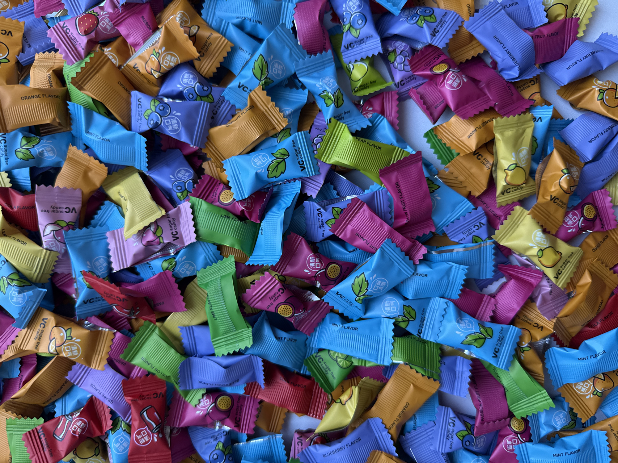 The image shows a large, disorganized pile of individually wrapped candies. The candies are of various colors, including orange, blue, purple, pink, yellow, red, and green. Each candy wrapper features the text "VC sugar free candy" and an illustration of the fruit flavor contained within: orange, blueberry, grape, strawberry, lemon, lime, pear, watermelon, passion fruit, and cola. Text in a different language is also visible on the wrappers. The candies are scattered randomly across a white surface.